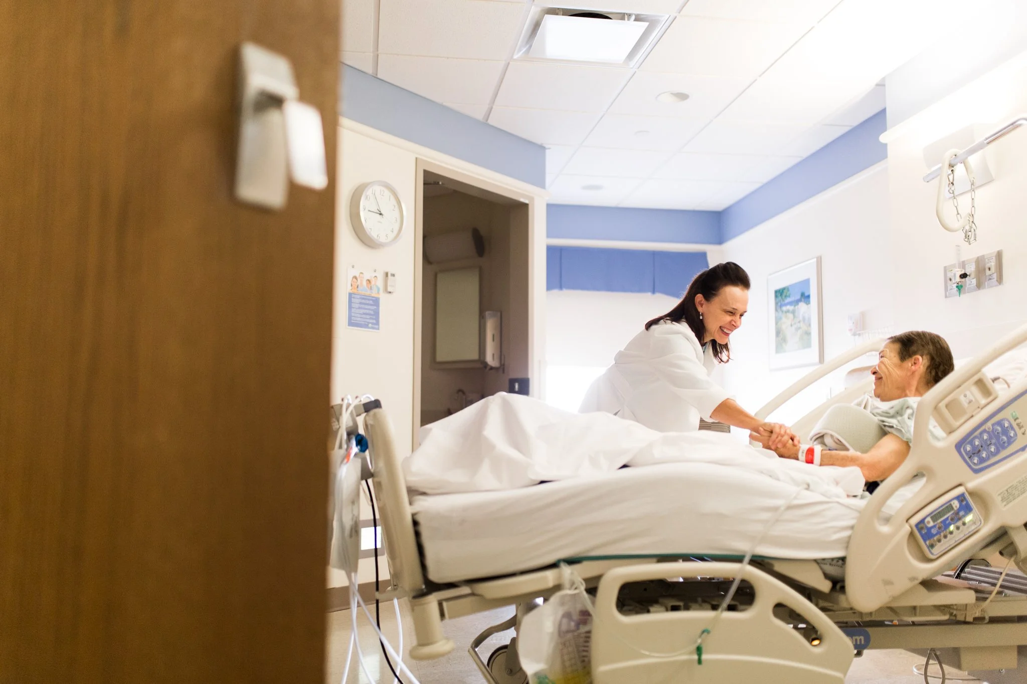 A St Elizabeth physician is smiling while holding hands with a patient lying in a hospital bed in a hospital room.