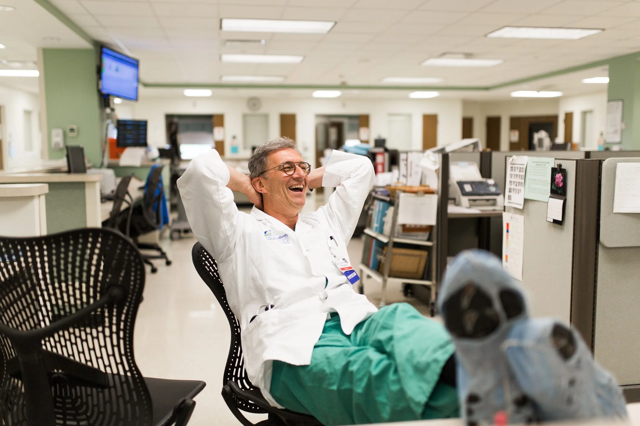 A male healthcare worker in green scrubs and a white coat is sitting in a chair in a hospital or medical facility, smiling and relaxing with arms behind his head. The background features cubicles, medical equipment, and monitors.