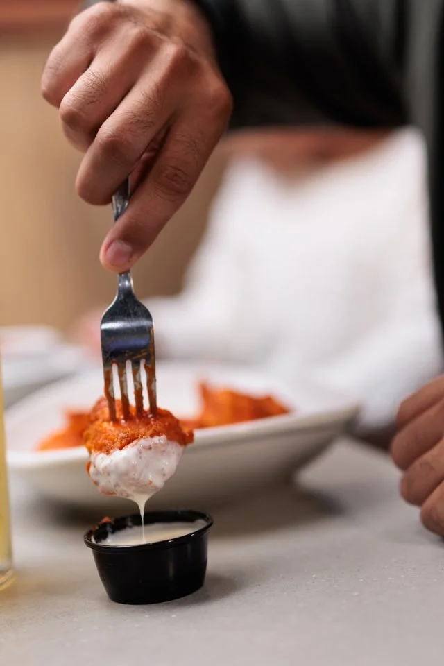 Person using a fork to dip a breaded piece of food into a white creamy sauce from a small black cup on a table.