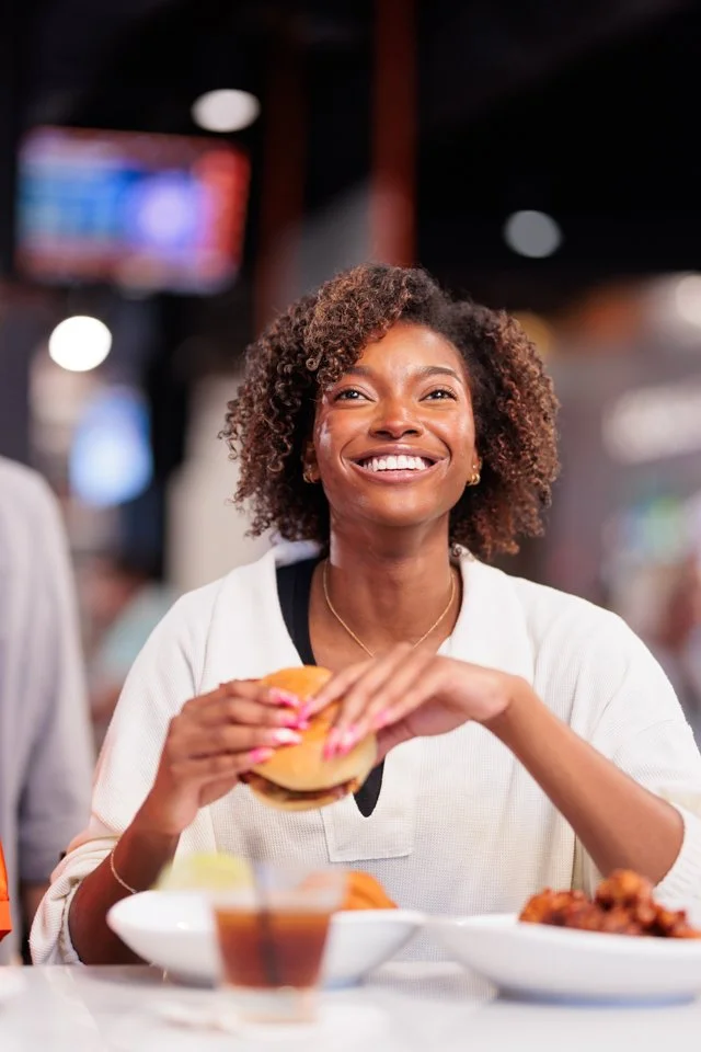 A smiling woman in a white shirt holding a burger at a restaurant table.