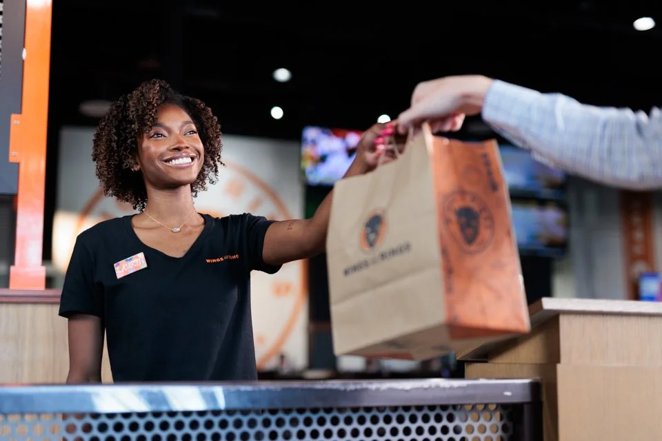 A happy female employee in a black uniform hands a takeout bag to a customer at a fast-food restaurant counter.