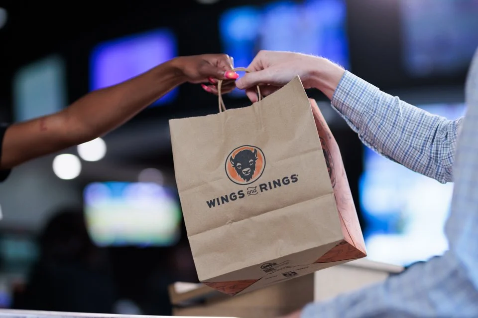 Person handing a paper bag labeled "Wings and Rings" to another person in a setting with multiple screens in the background.