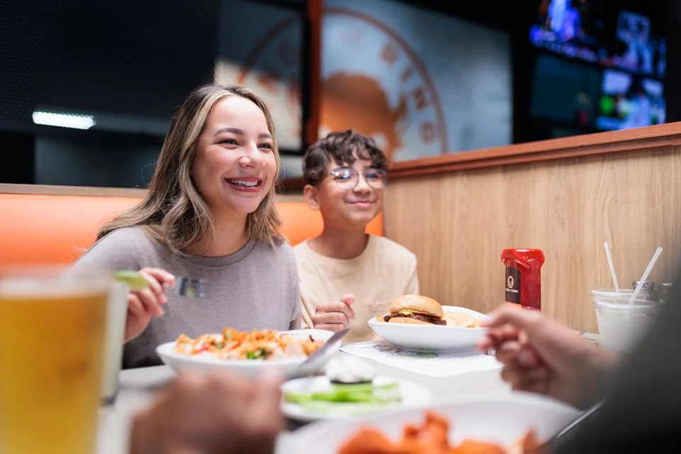 Two young adults, a woman and a man, smiling and enjoying a meal at a casual restaurant. The woman is holding utensils with a bowl of salad in front of her, and the man has a burger on his plate. There are drinks, ketchup, and side dishes on the tabl