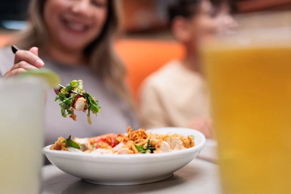 A woman is smiling and holding a forkful of food over a bowl of fried rice in a restaurant. There is a large glass of yellow beverage in the foreground.