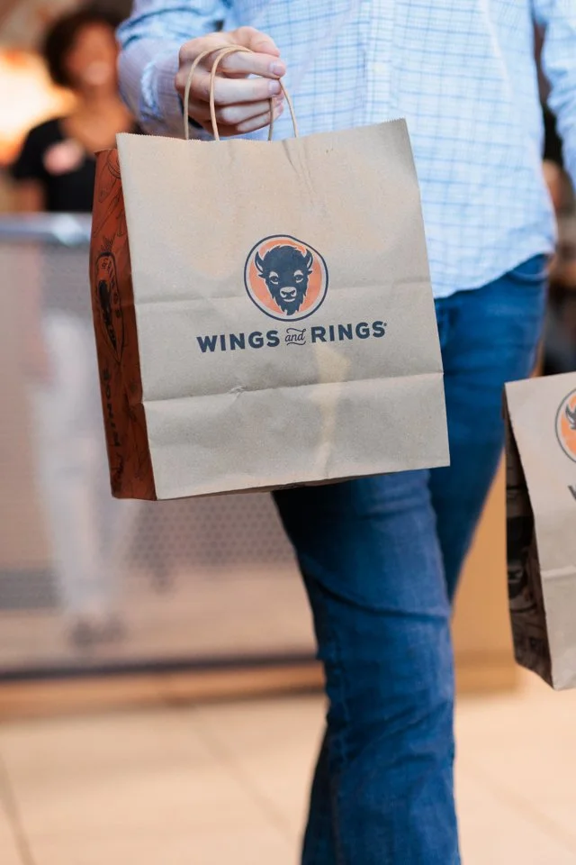 Person holding a paper bag with the Wings and Rings logo, a cartoonish buffalo face, in a casual setting.