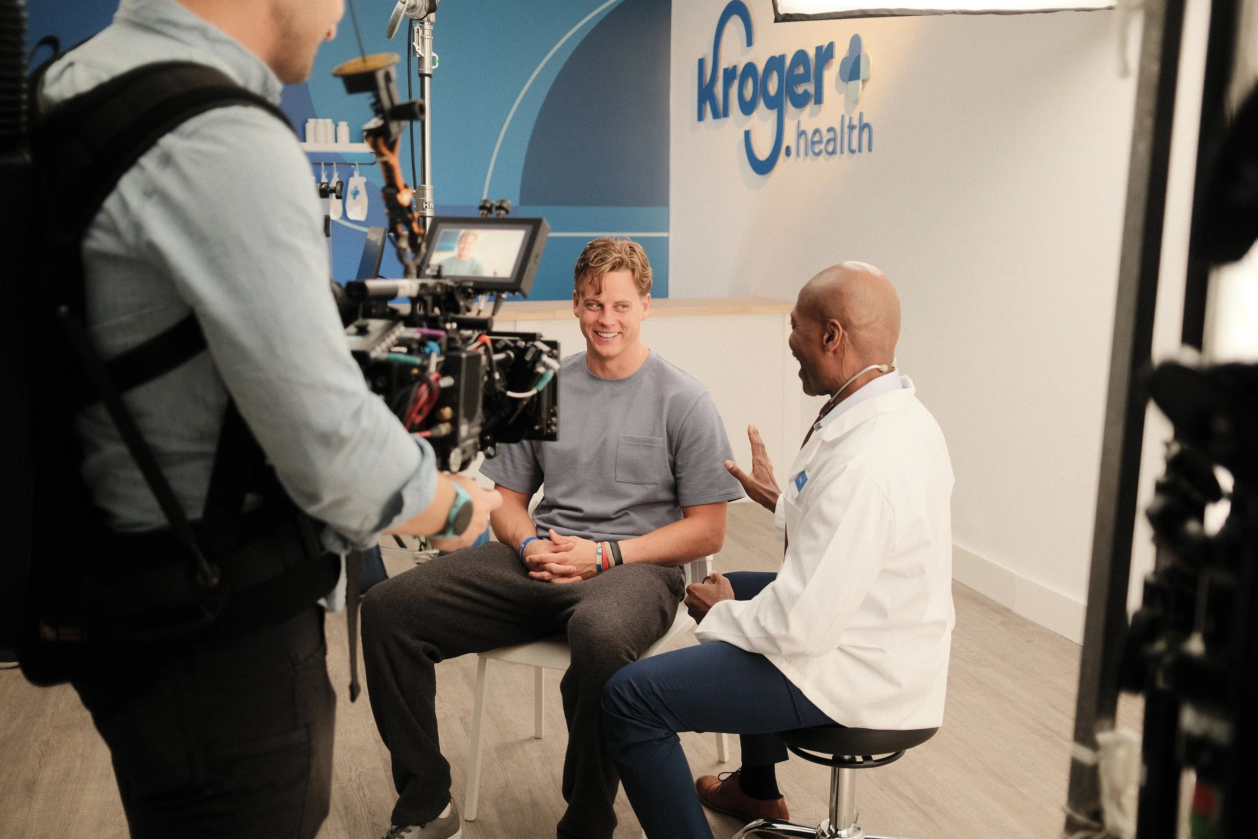 A young man is sitting on a white chair in a medical setting, talking to a doctor. The doctor is engaged in a conversation, using hand gestures. A camera crew is filming the scene, with a cameraman operating a large camera. The background features a 