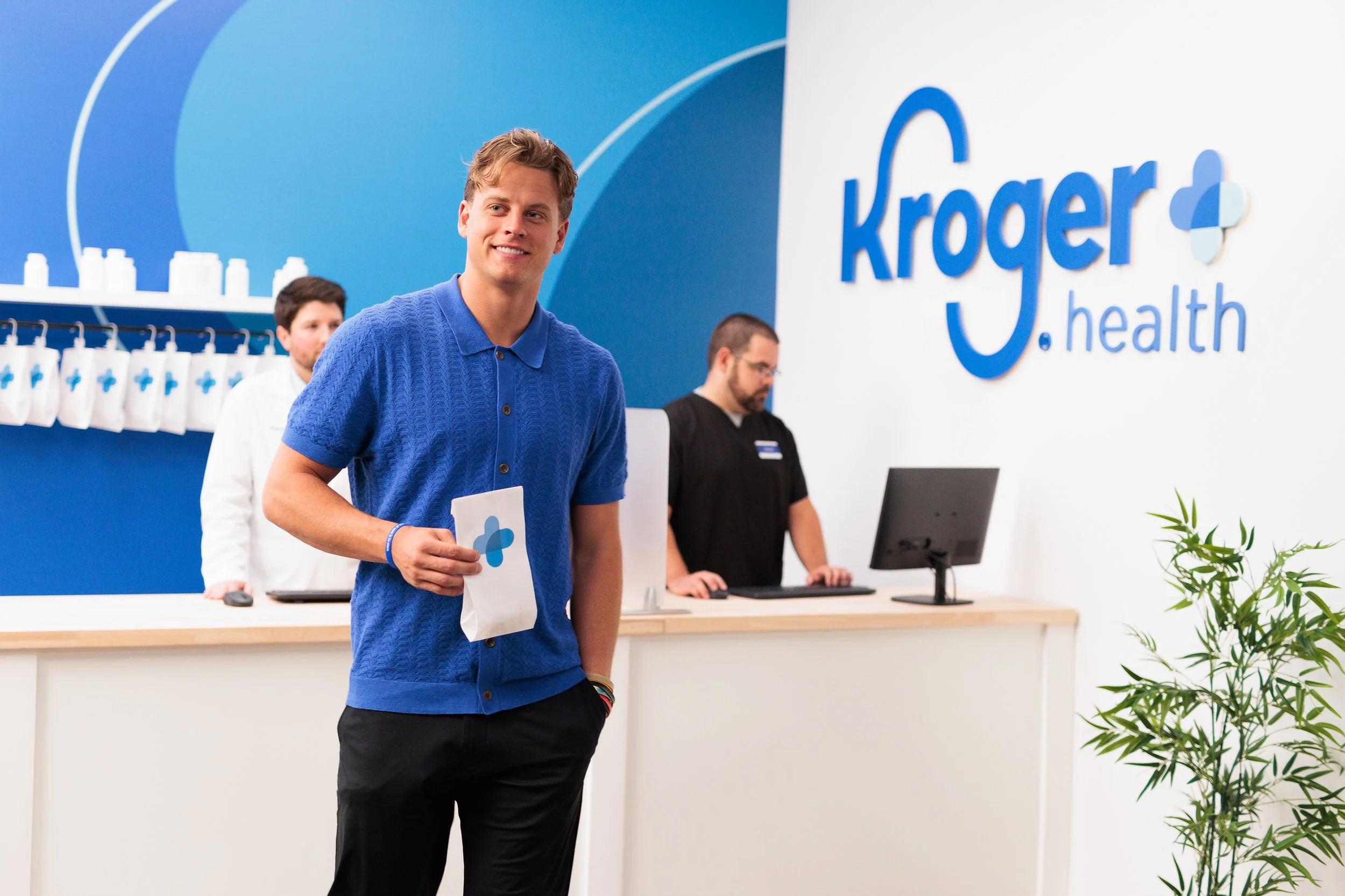 Joe Burrow is holding a Kroger Health shopping bag standing at a reception desk in a Kroger Health store, with two employees working behind the counter in a modern, bright interior.