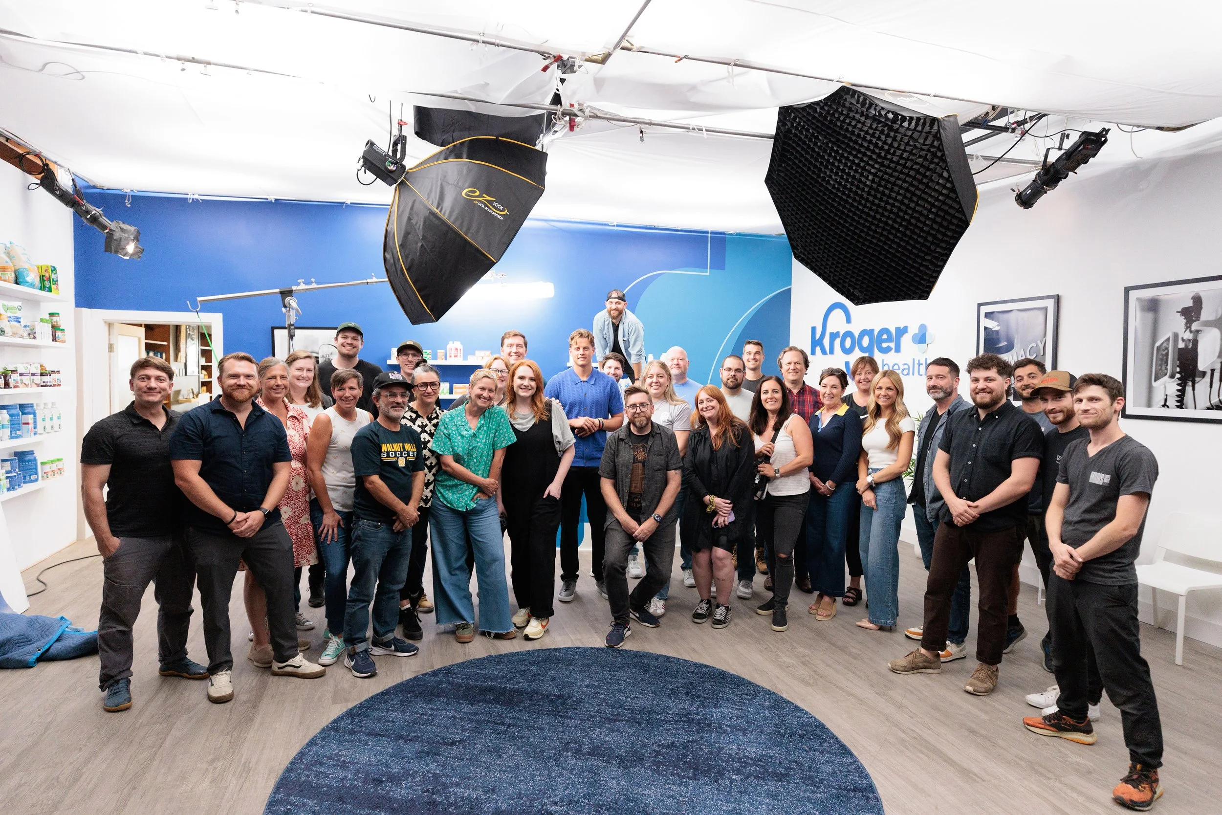 Group of people posing in a studio with Kroger Health logo on the wall, photography equipment overhead, shelves with supplies, and framed pictures on the wall.