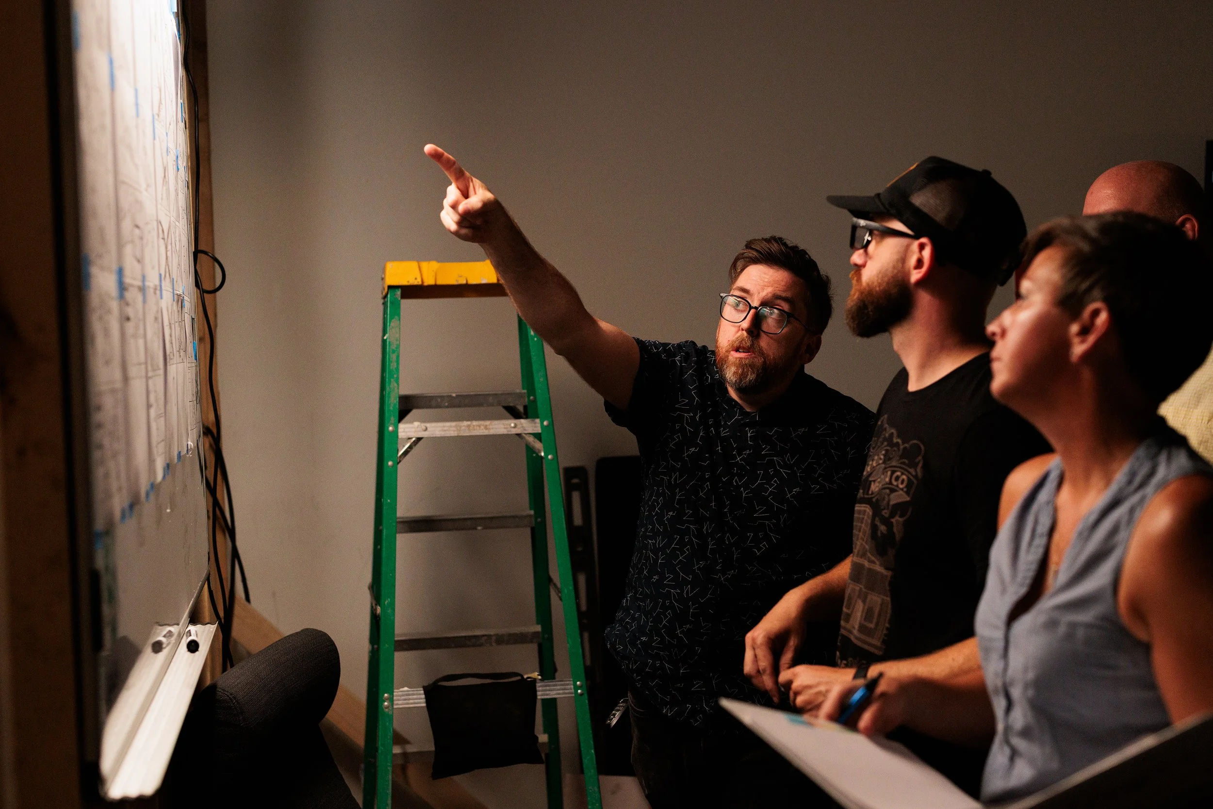 A man with glasses and a beard is pointing at a whiteboard while explaining to a group of three people in a room with a ladder behind him.