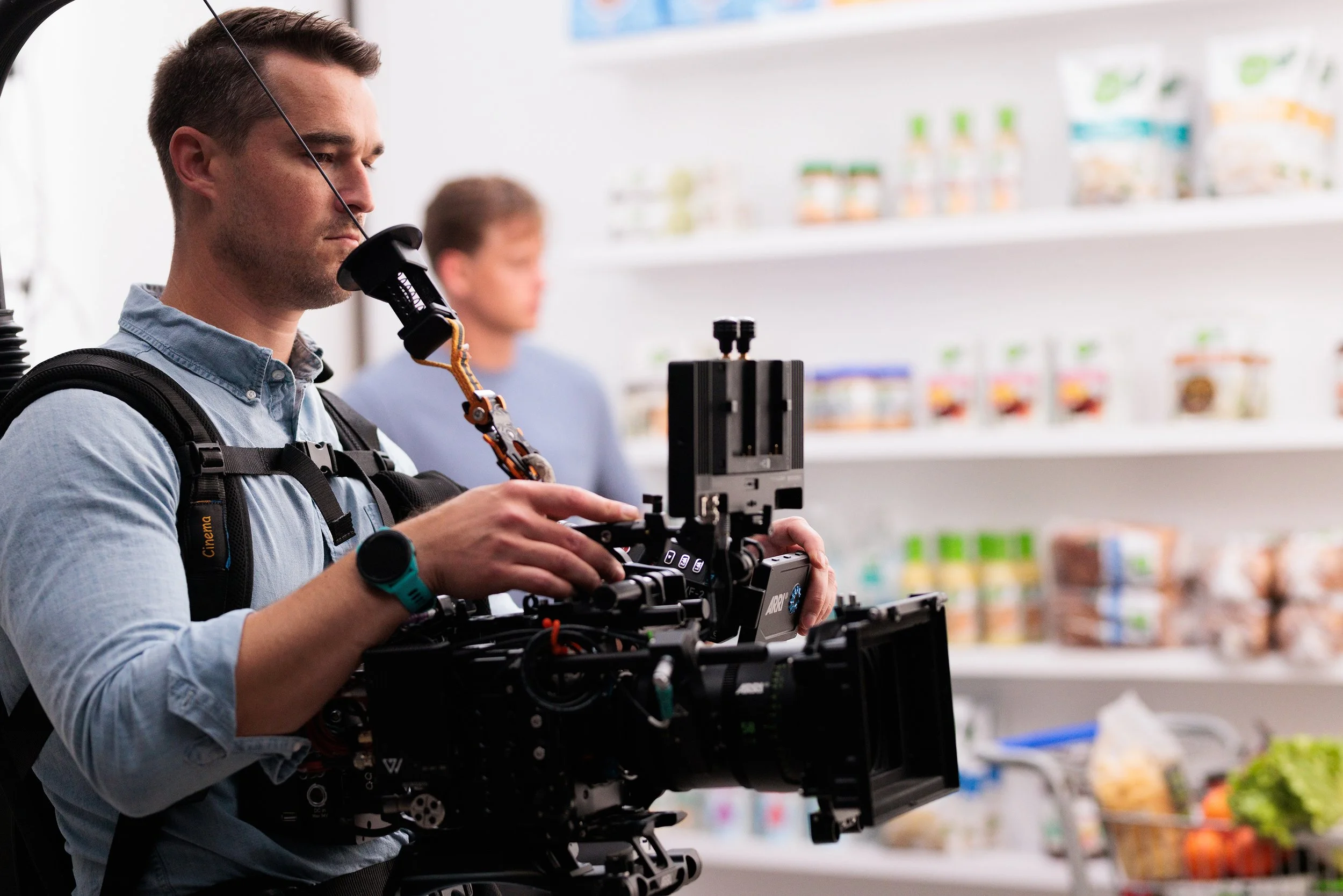 A man filming with a professional camera in a grocery store, with a blurred Joe Burrow shopping in the background.