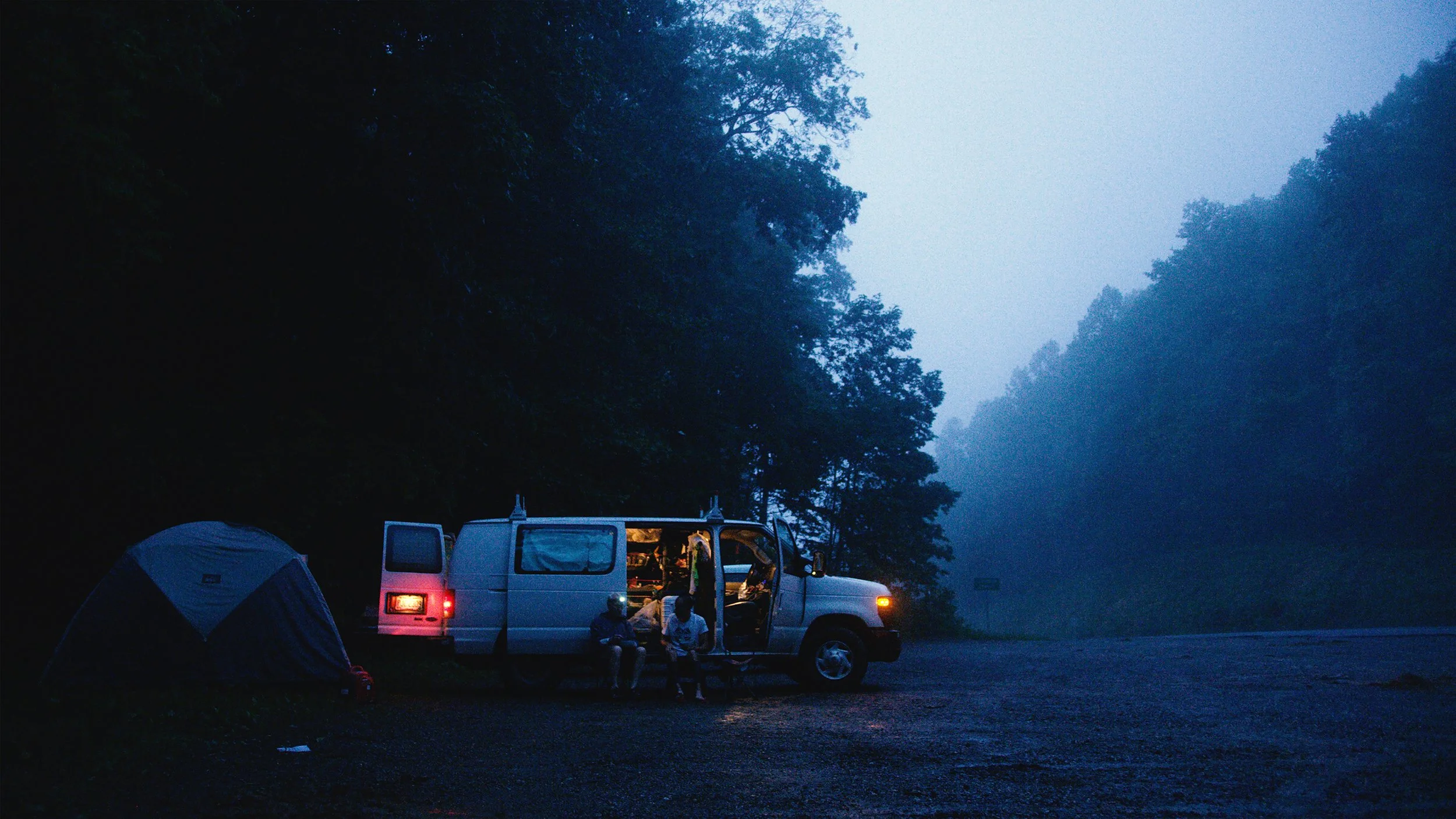 A nighttime camping scene with a white van with open door, two people sitting outside, a tent, and a forested background with fog.