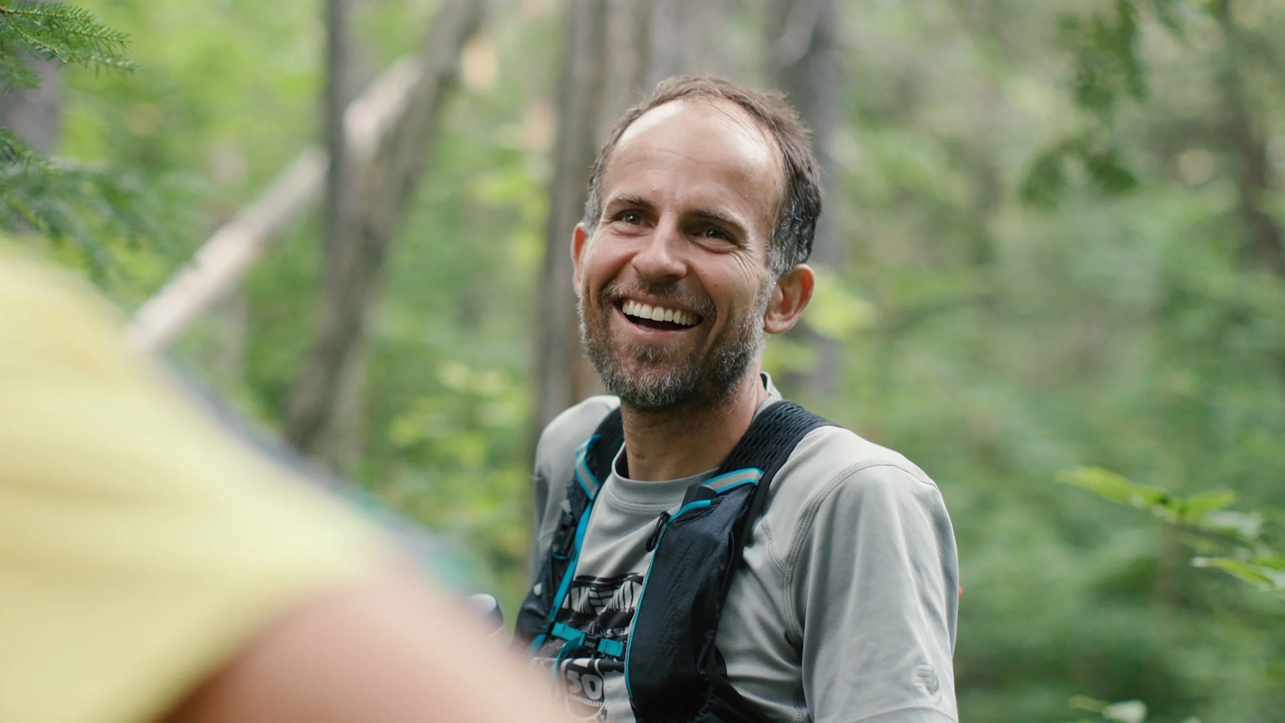 Harvey Lewis smiling while trail running in the woods, wearing a grey shirt and a black backpack.