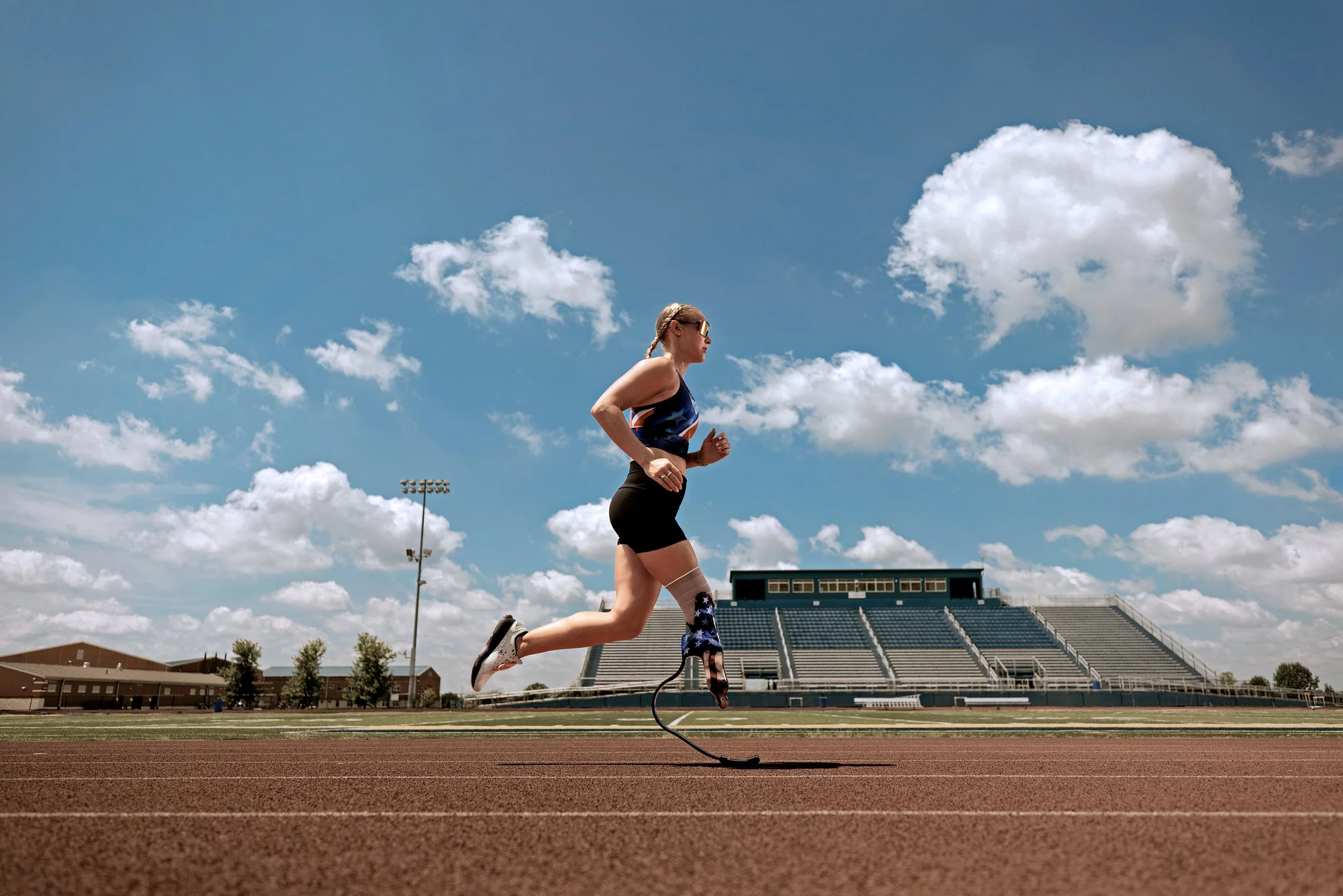 A female runner with a prosthetic leg running on a track field under a partly cloudy sky.