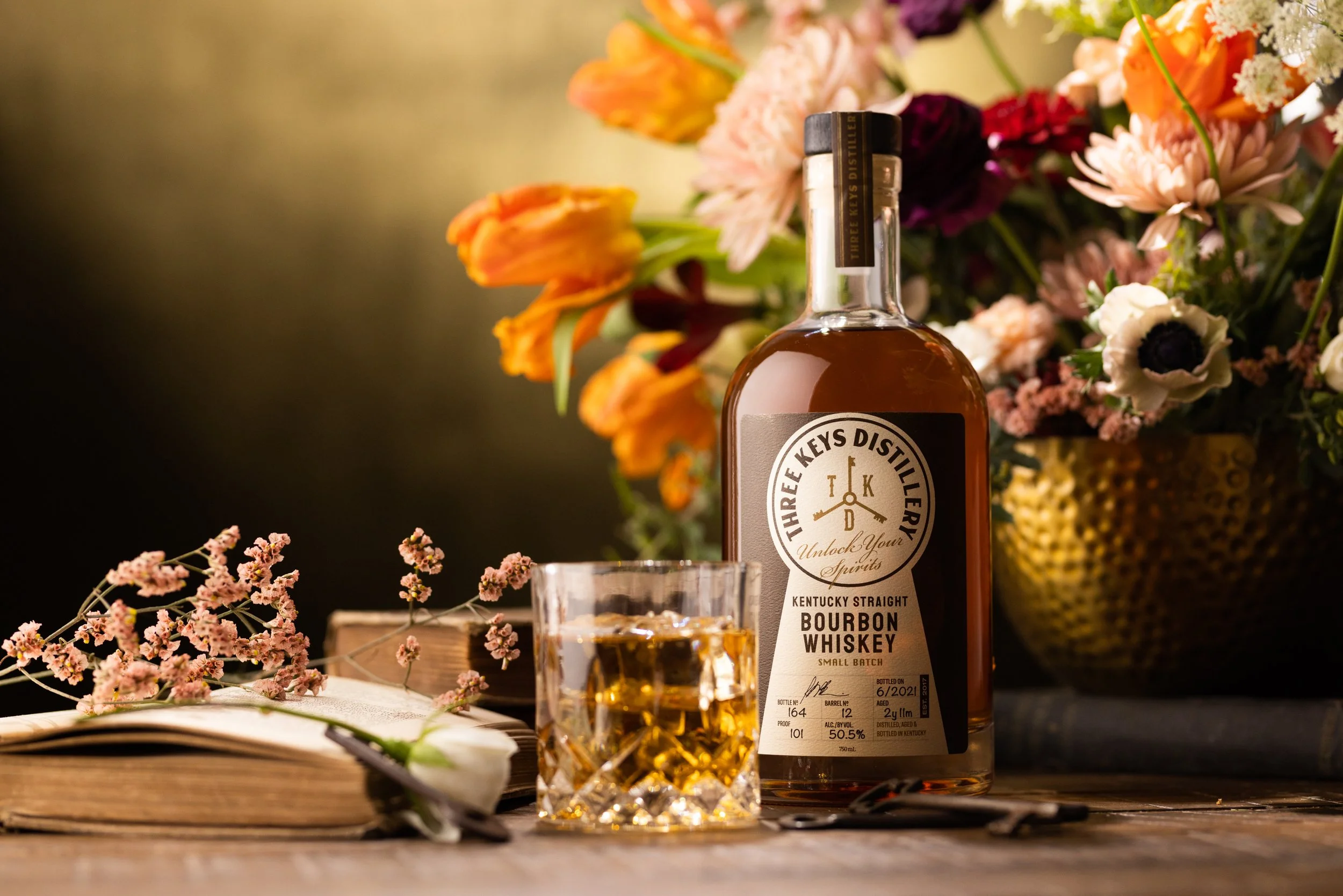 A bottle of Three Keys Distillery Kentucky Straight Bourbon Whiskey, a glass filled with whiskey, a flower, an open book, and a bouquet of colorful flowers in a gold vase on a wooden surface.