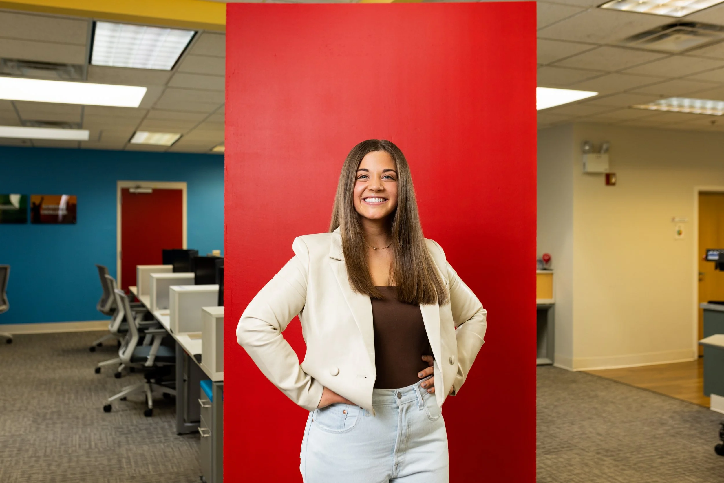 Smiling young woman with long brown hair, wearing a white blazer over a dark top and light jeans, standing in front of a red wall in an office setting.