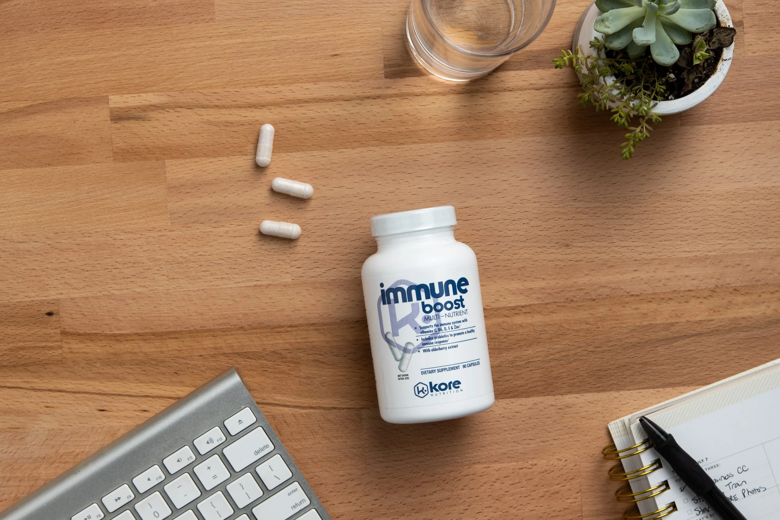 A white bottle labeled 'immune boost' with capsules nearby, a glass, a potted succulent plant, a keyboard, a notebook with a pen, all on a wooden desk.