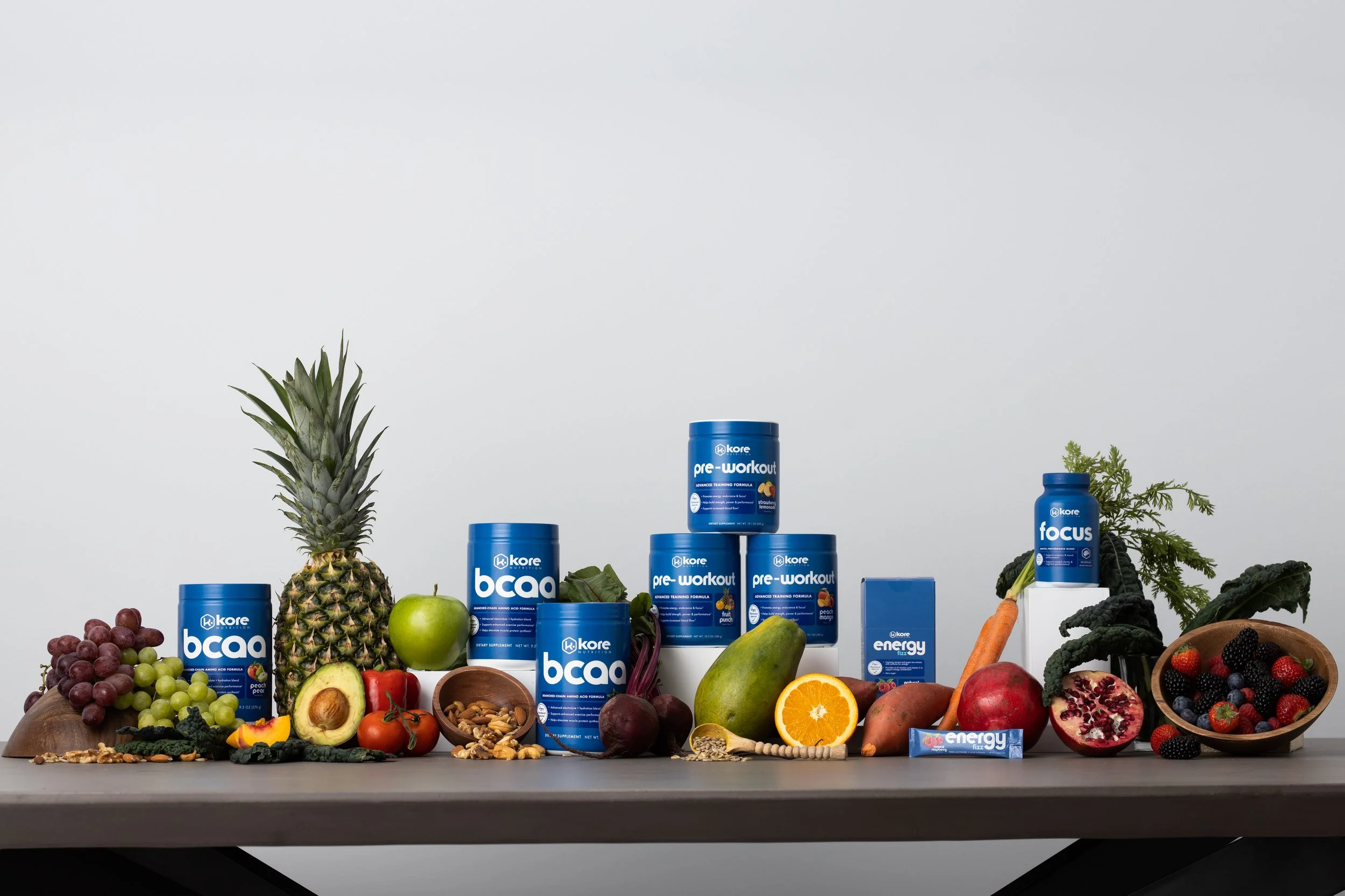 An arrangement of dietary supplements and fresh fruits on a table. The supplements include powders labeled 'bca,' 'pre-workout,' 'energy,' and 'focus.' The fruits include pineapple, green apple, avocado, orange, beet, mango, pomegranate, grapes, carr