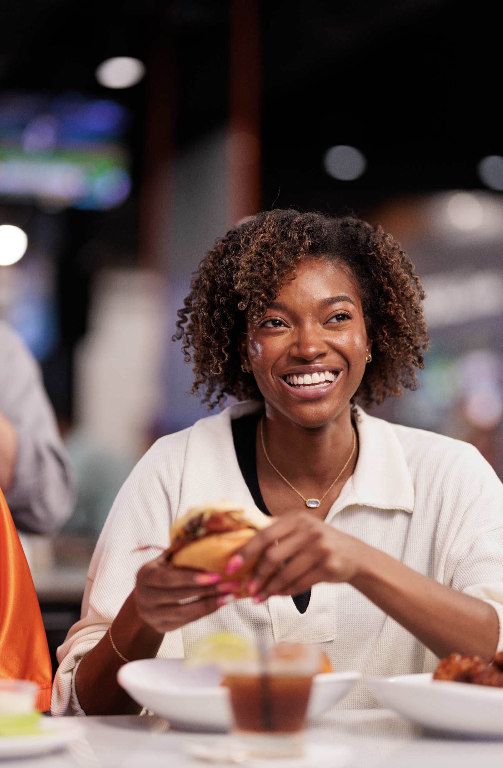A woman with curly hair smiling while holding a sandwich in a restaurant or cafe setting.
