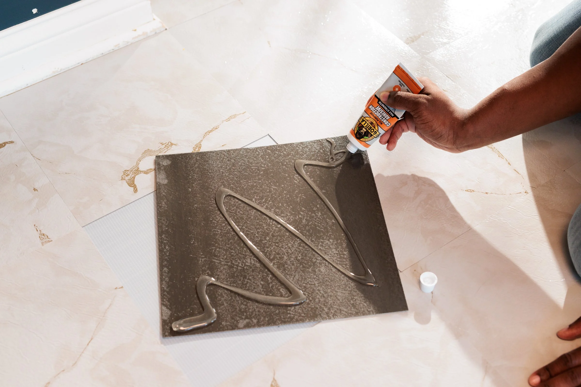 A person is applying glue from a tube onto a tile with a decorative pattern, creating a zigzag design.