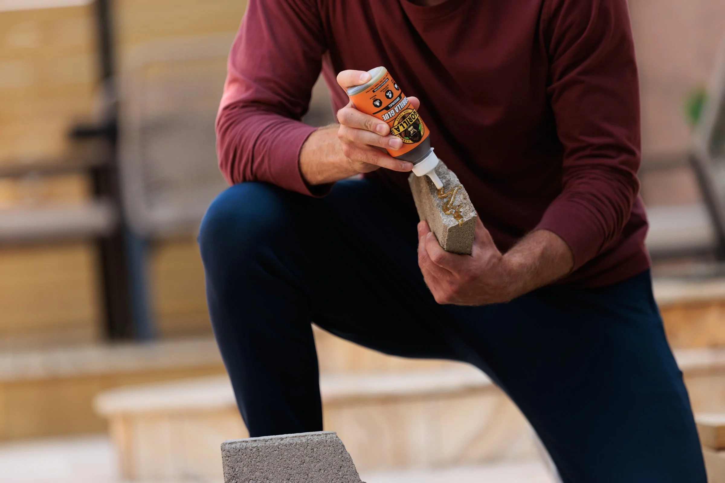 A person kneeling outdoors, applying construction adhesive from a caulking tube to a concrete brick.