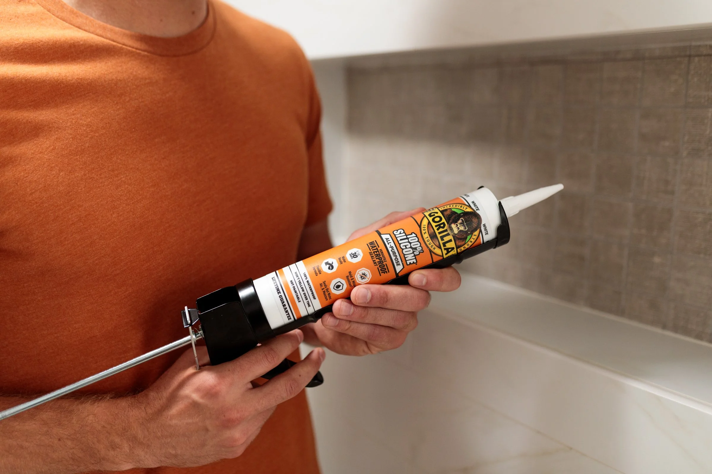 A person in an orange shirt holding a tube of Gorilla 100% Silicone Sealant with a white nozzle, preparing to apply it to a surface.