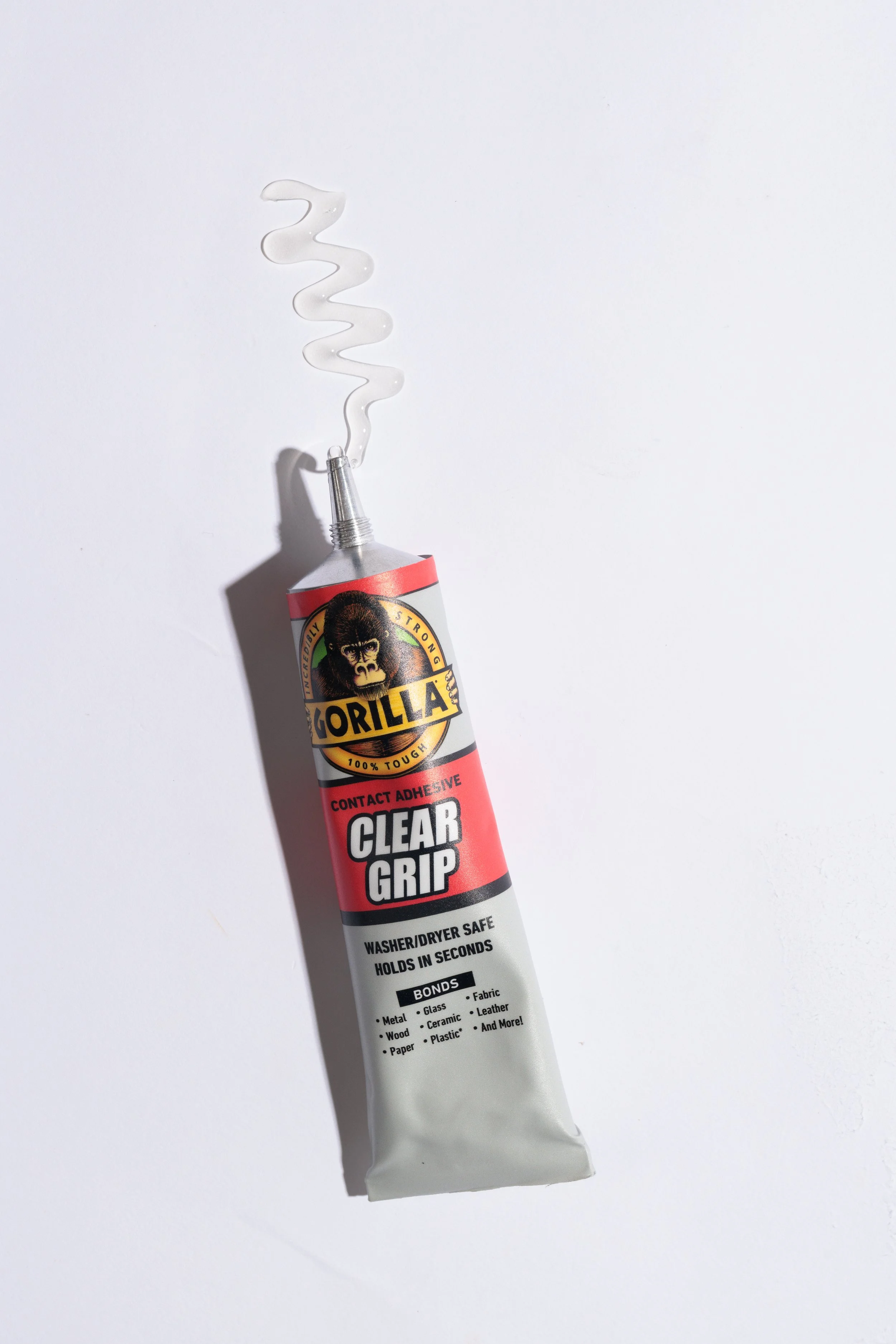 A tube of Gorilla brand clear grip contact adhesive with a small amount of white glue coming out of its nozzle, placed on a white surface.