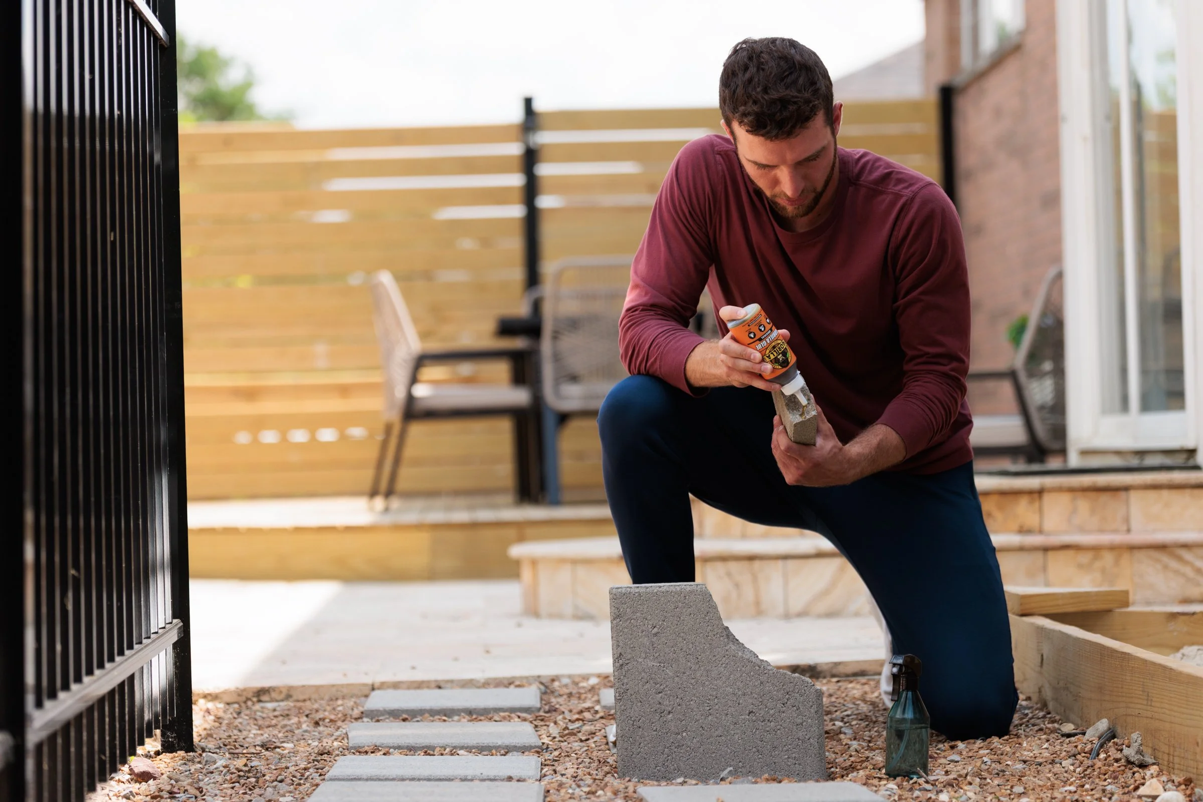 Man kneeling outdoors, applying adhesive to a cinder block for a home improvement project.