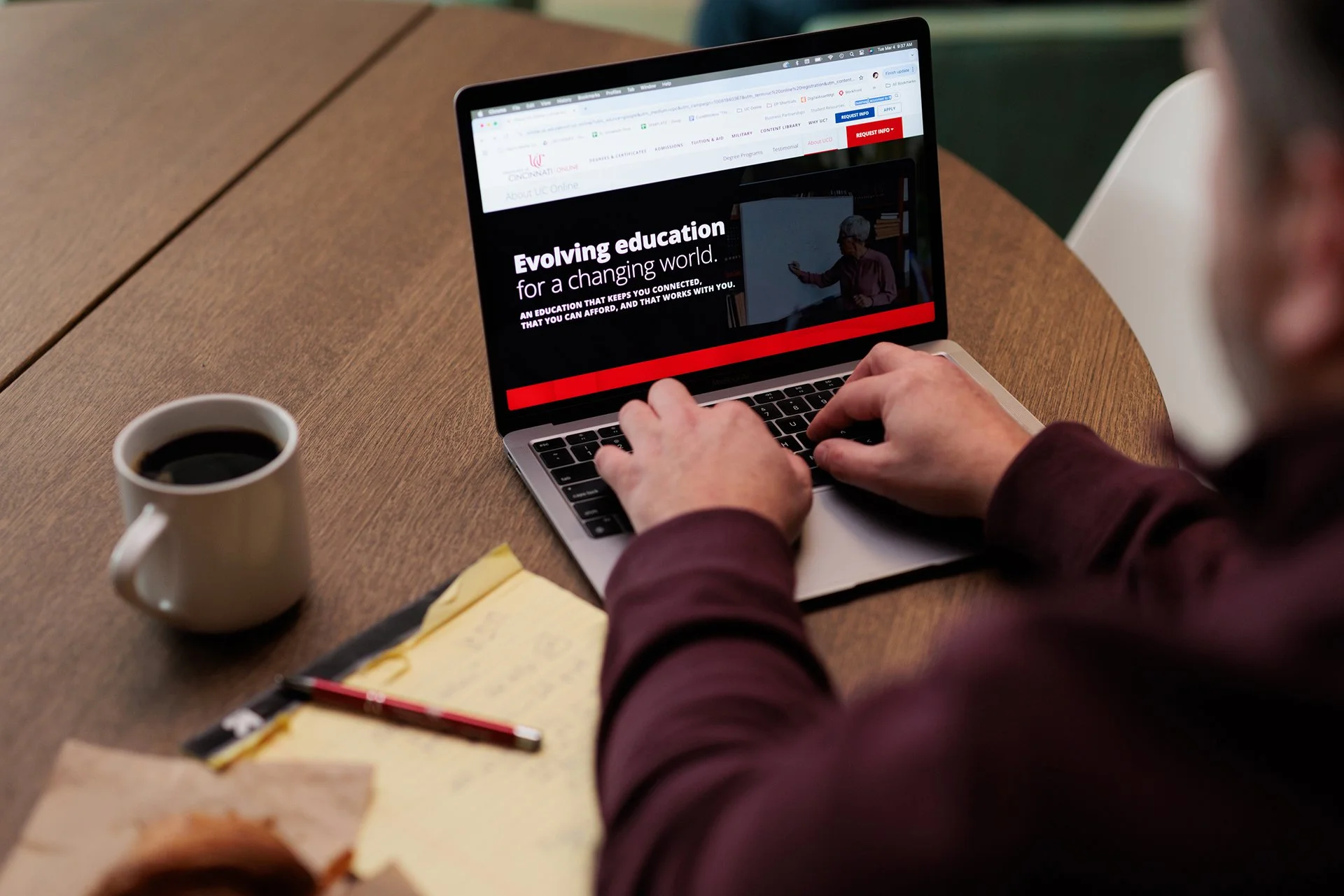 Person using a laptop with a webpage about evolving education on a wooden table, next to a cup of coffee, a yellow notepad, and a pen.
