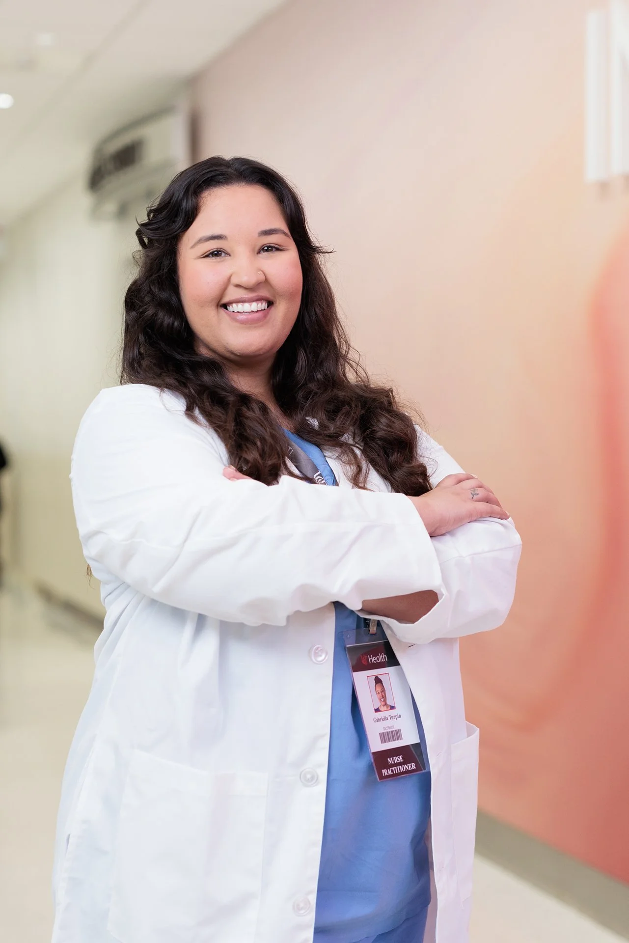 Smiling female nurse with long dark curly hair standing with arms crossed in a hospital corridor, wearing a white coat and navy blue scrubs with a name badge.