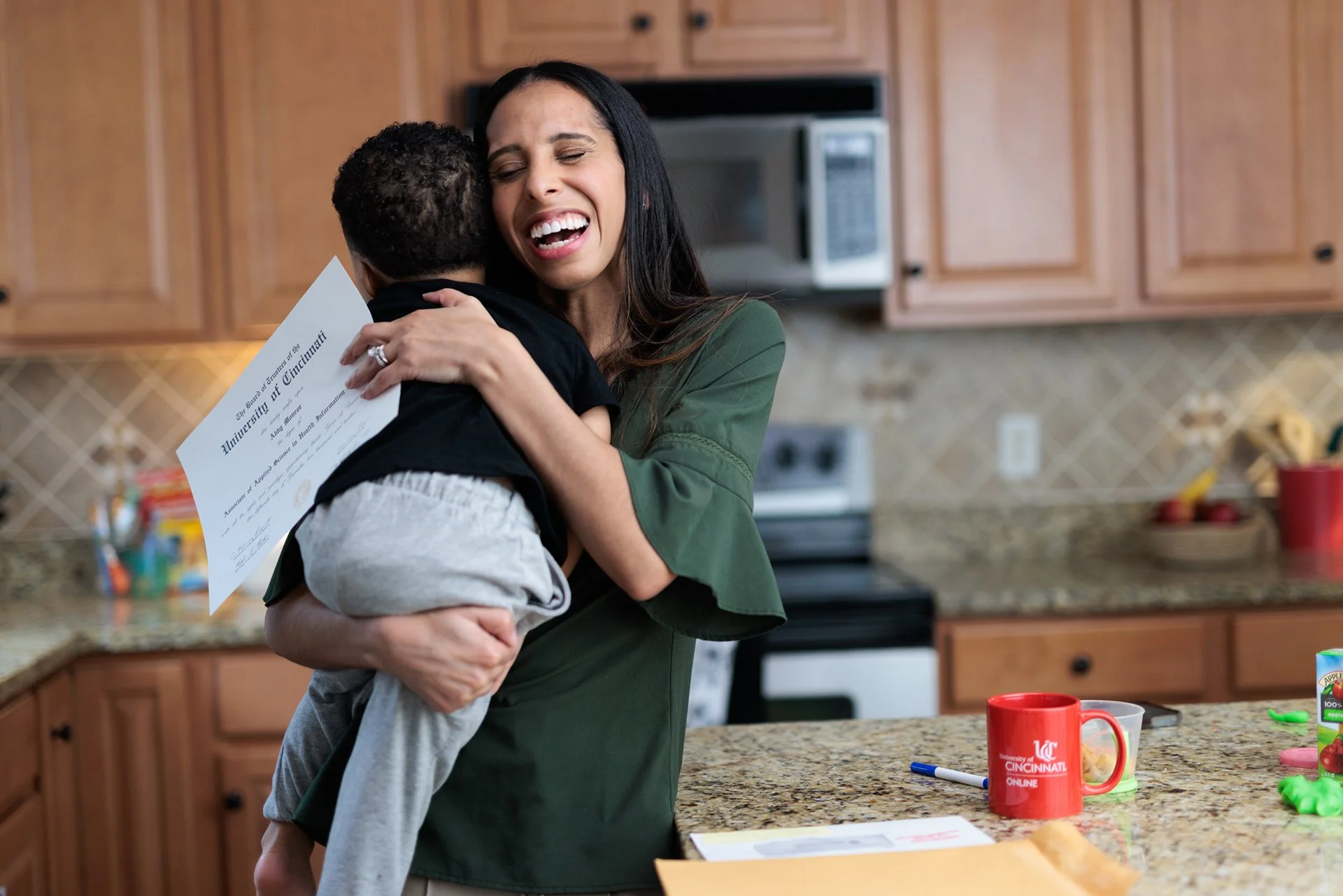 Woman hugging a child while holding a diploma in a kitchen