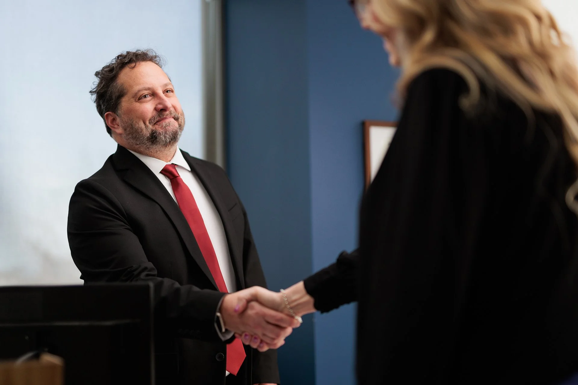 A man in a black suit with a red tie shaking hands with a woman in a black outfit in an office setting.