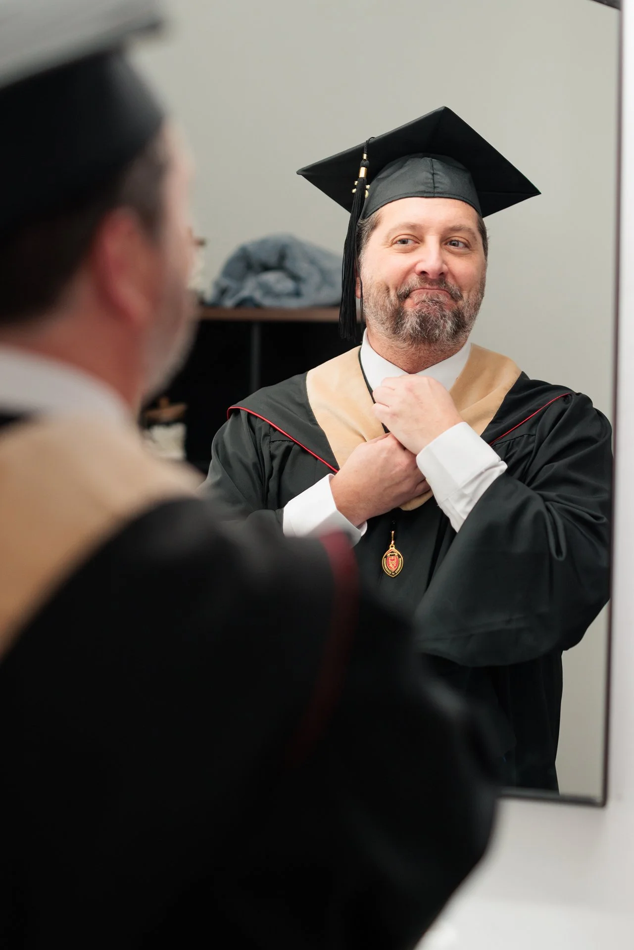 A man wearing graduation cap and gown looking at himself in a mirror, adjusting his gown with a smile