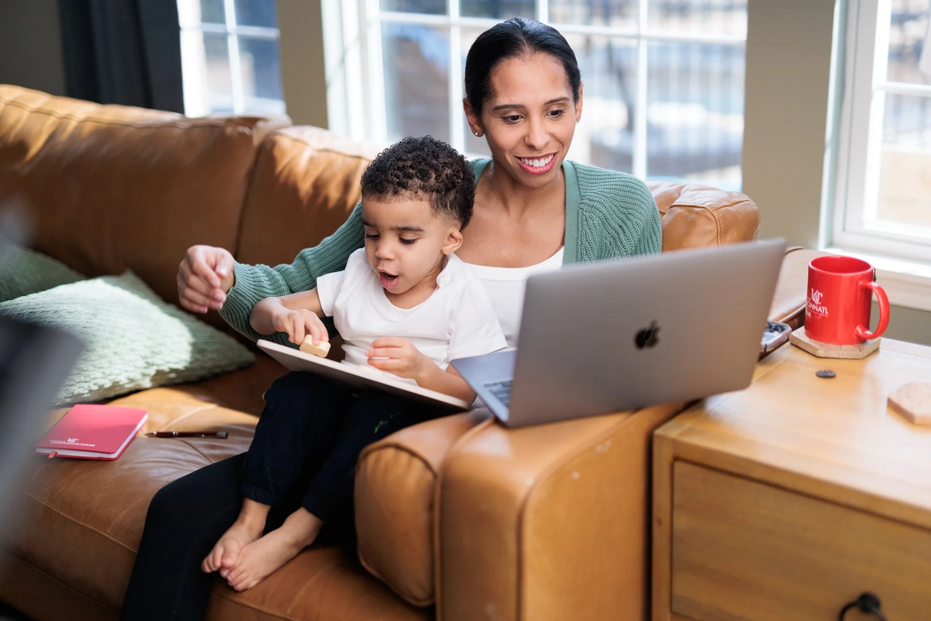 A woman and young boy sit together on a leather couch in a brightly lit room, with the woman smiling as the boy looks at a tablet in his hands, while a laptop and a red mug are on a side table.