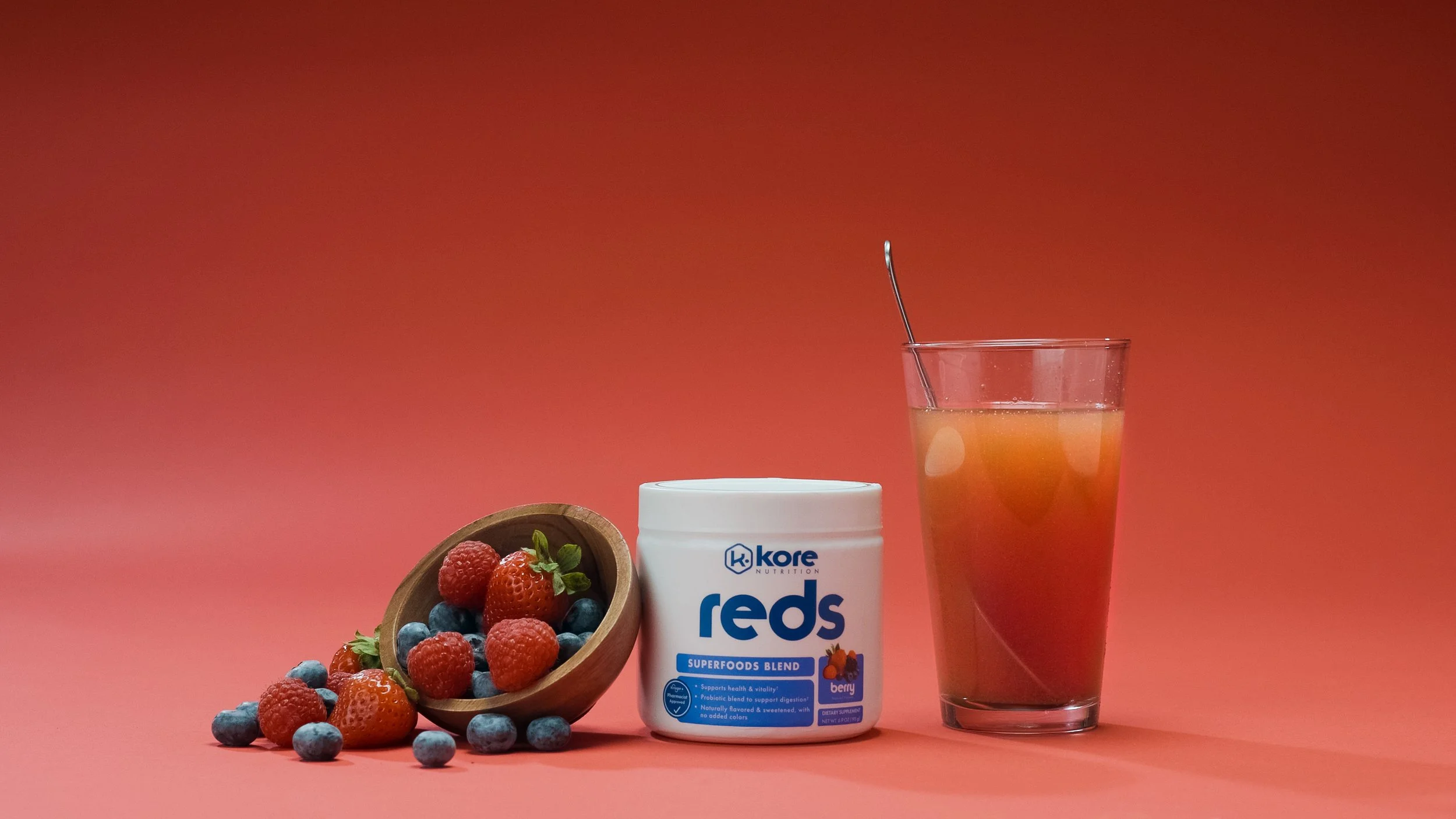 A container of Kore Reds berry superfood powder, a glass of fruit juice with ice, and a wooden bowl filled with strawberries, raspberries, and blueberries against a pink background.