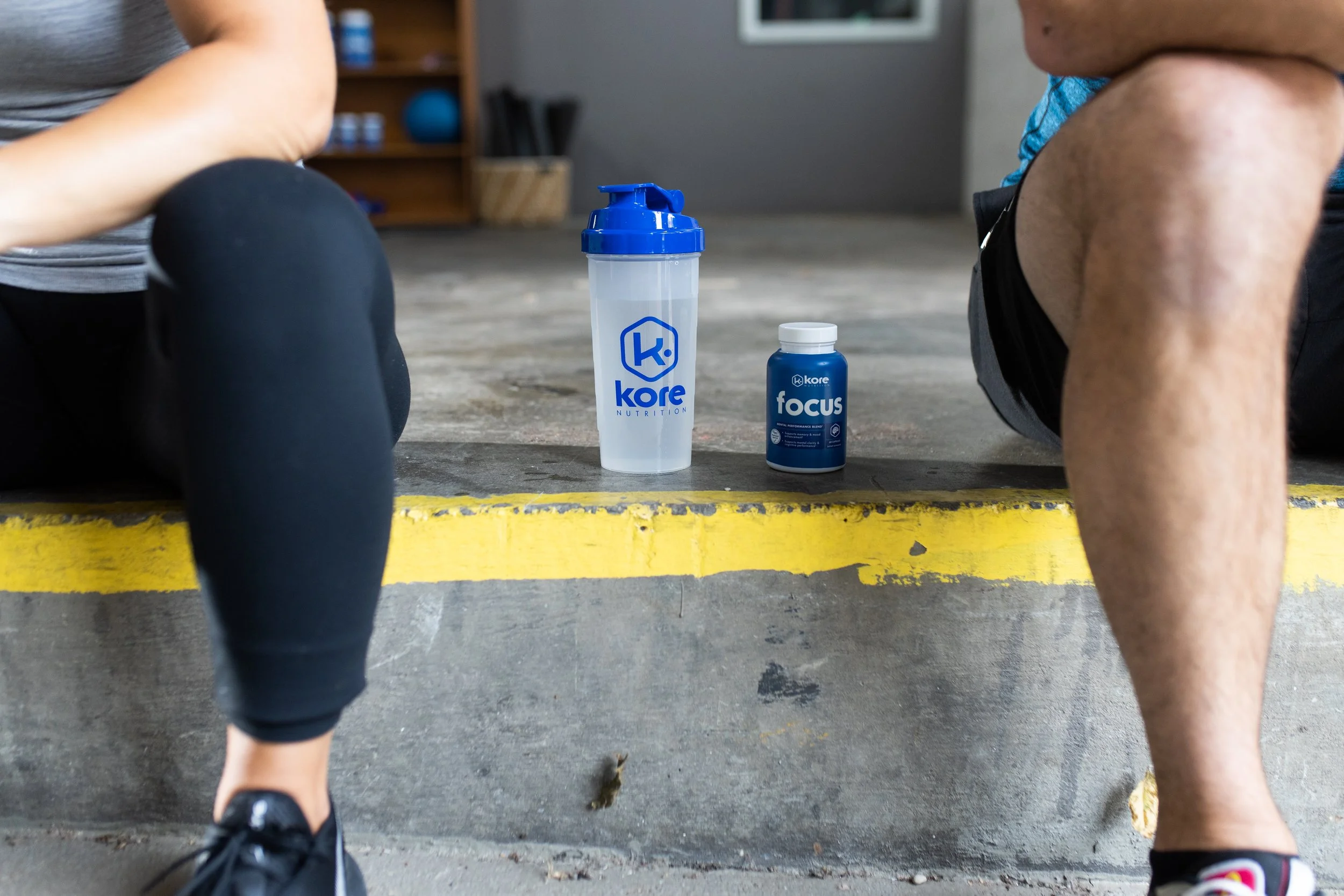 A close-up of two people sitting on a concrete curb, with fitness water bottles labeled 'Kore Nutrition' and 'Focus' placed between them. The background shows a gym or fitness studio setting.