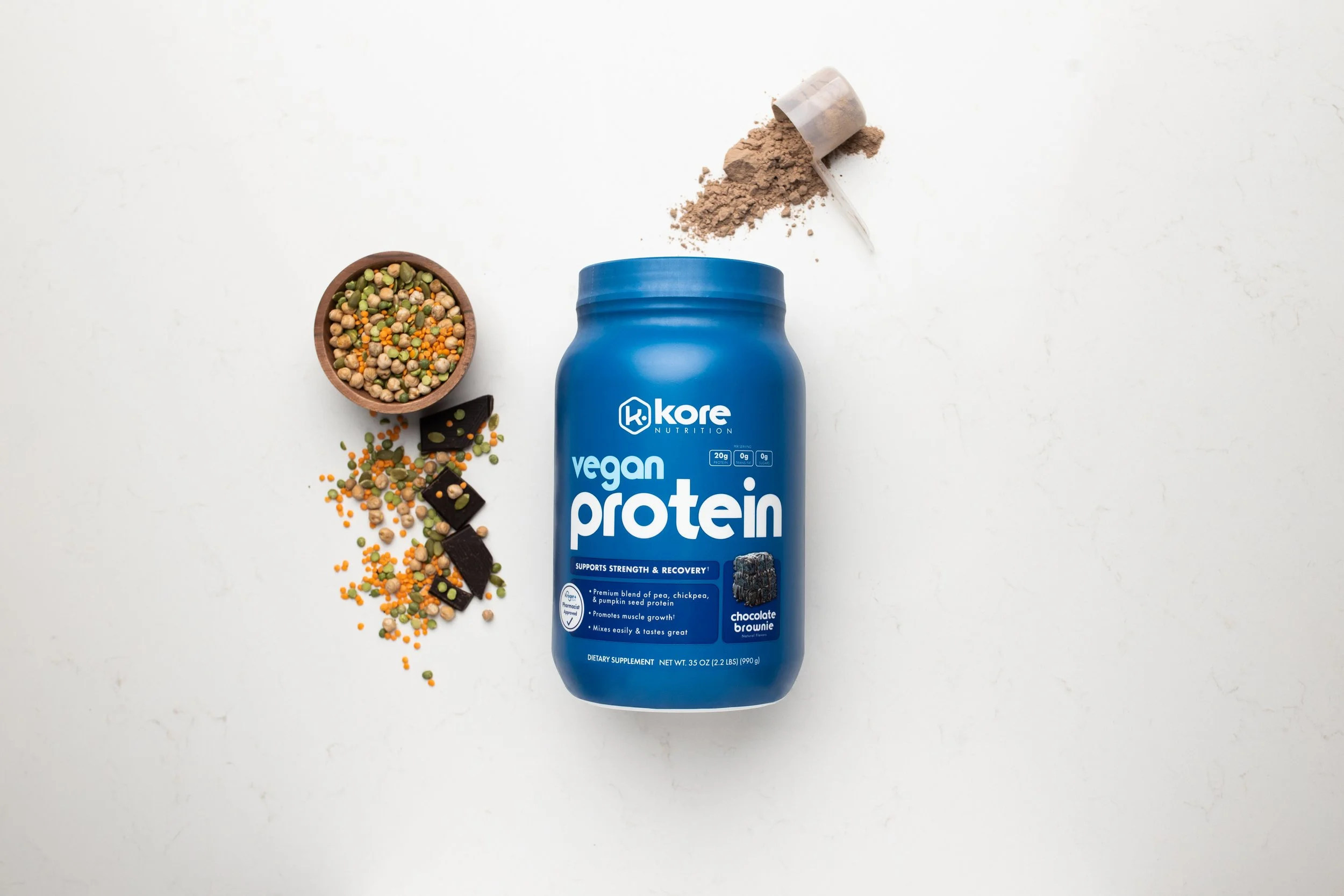 Blue container of vegan protein powder with scoops, lentils, chickpeas, pumpkin seeds, and chocolate brownie pieces on a white background.