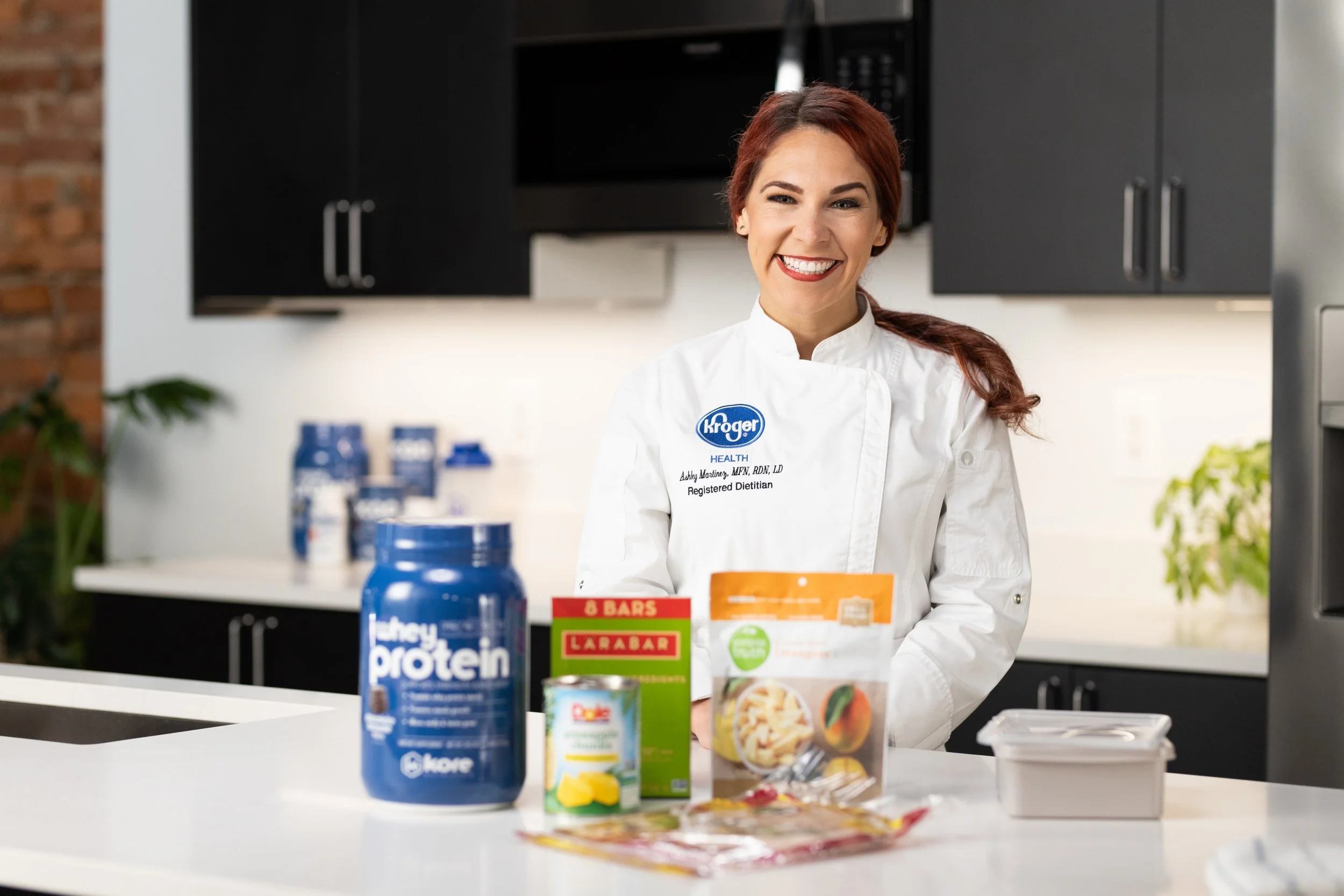 A smiling woman in a white chef's coat stands behind a kitchen counter with various food and supplement products, including protein powder, snack bars, and canned fruit.