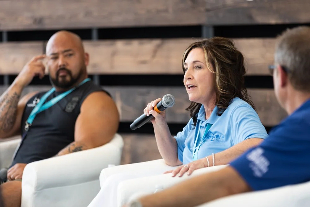 Woman speaking into a microphone during a panel discussion, with two men listening and one man appearing to be in thought.