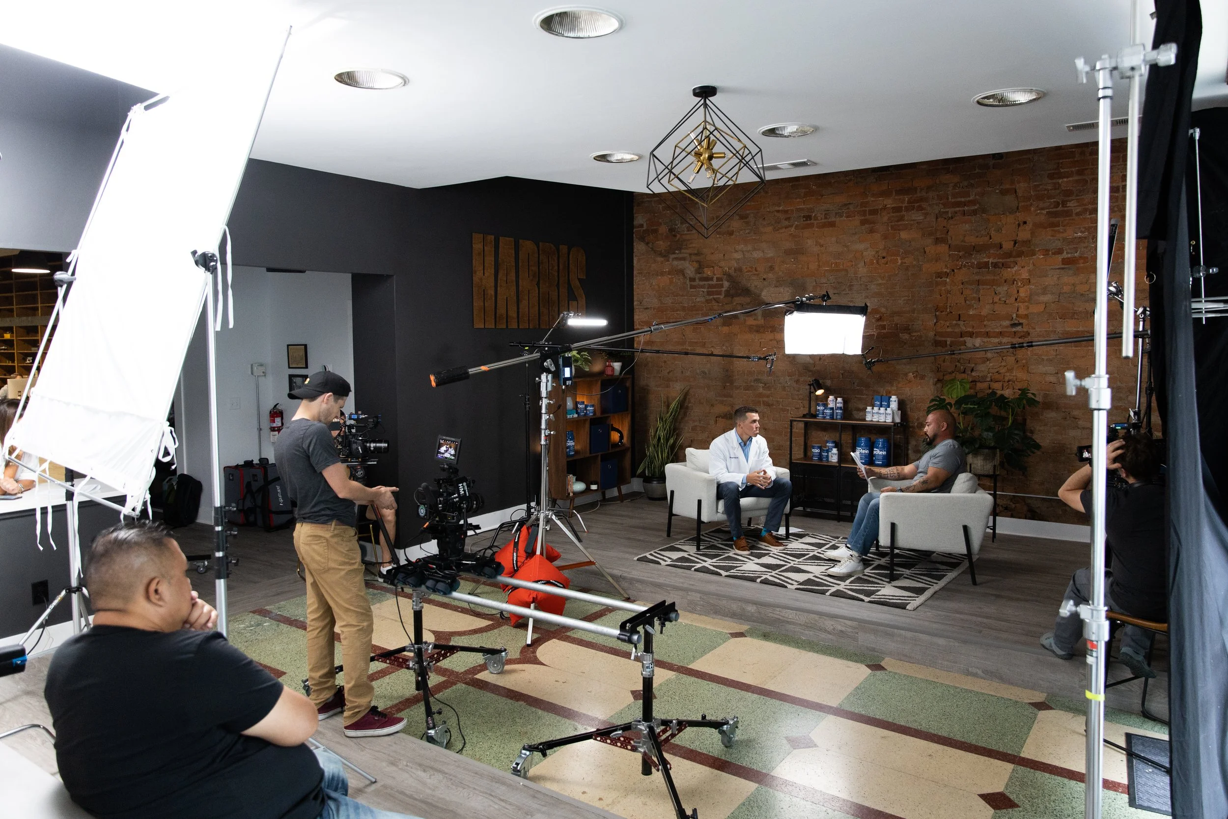 Two men sit on white armchairs in a studio setting with a brick wall background, during a filmed interview. One man wears a white coat, and the other is dressed casually. Camera crew and equipment, including cameras, lights, and boom microphones, sur