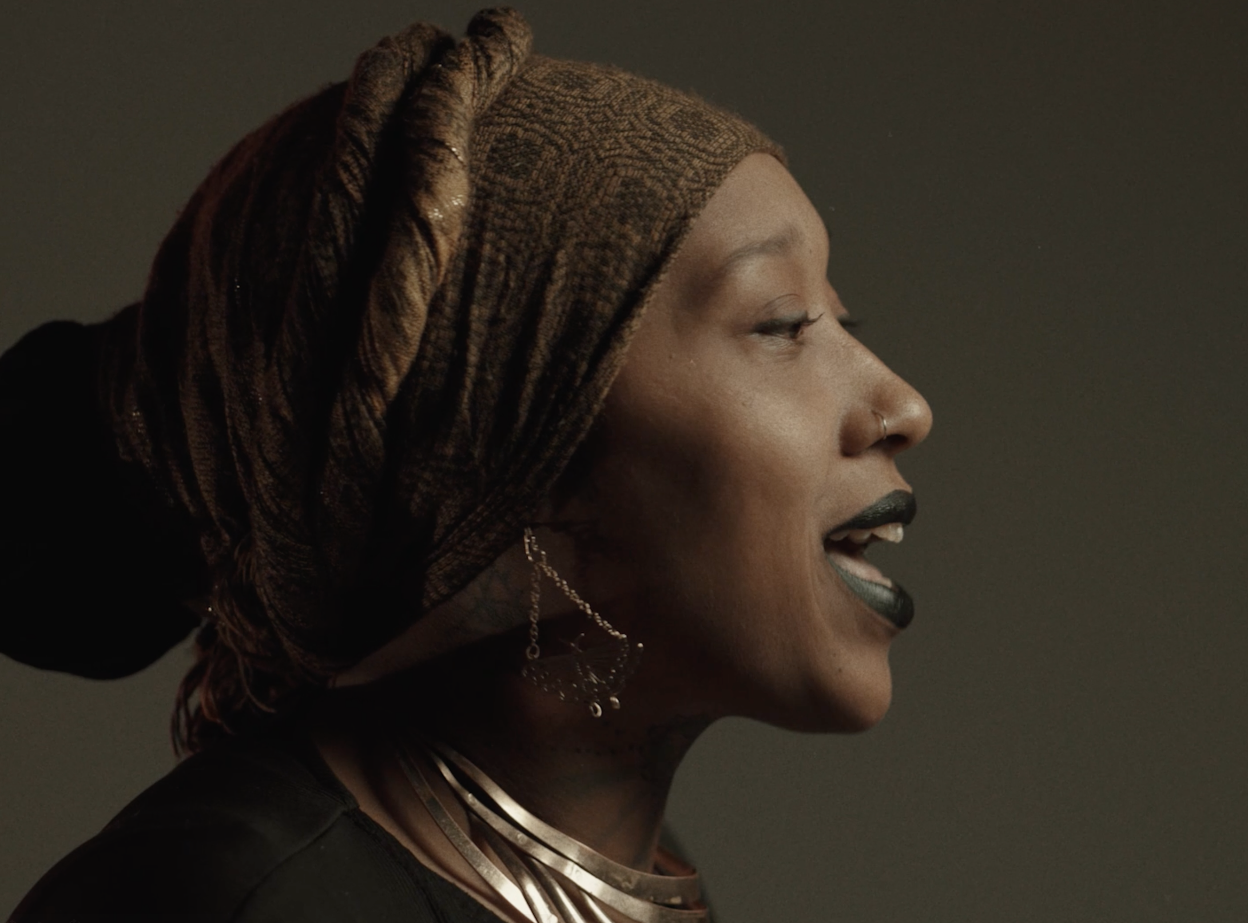 Side profile of a Black woman with braided headwrap, earrings, and jewelry, singing with eyes closed.