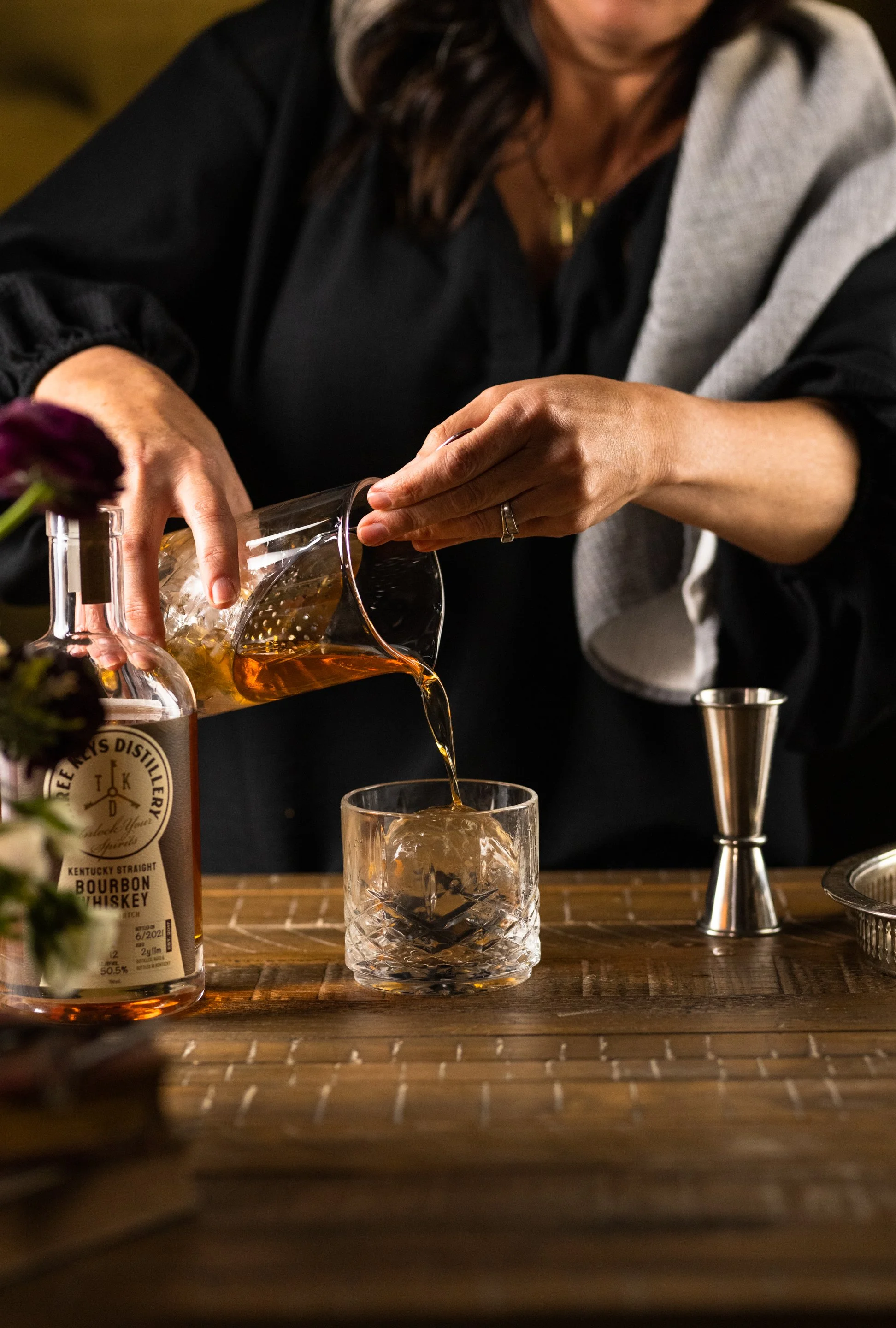 A person pours bourbon whiskey into a glass with ice on a wooden bar counter, with a bottle of bourbon nearby.
