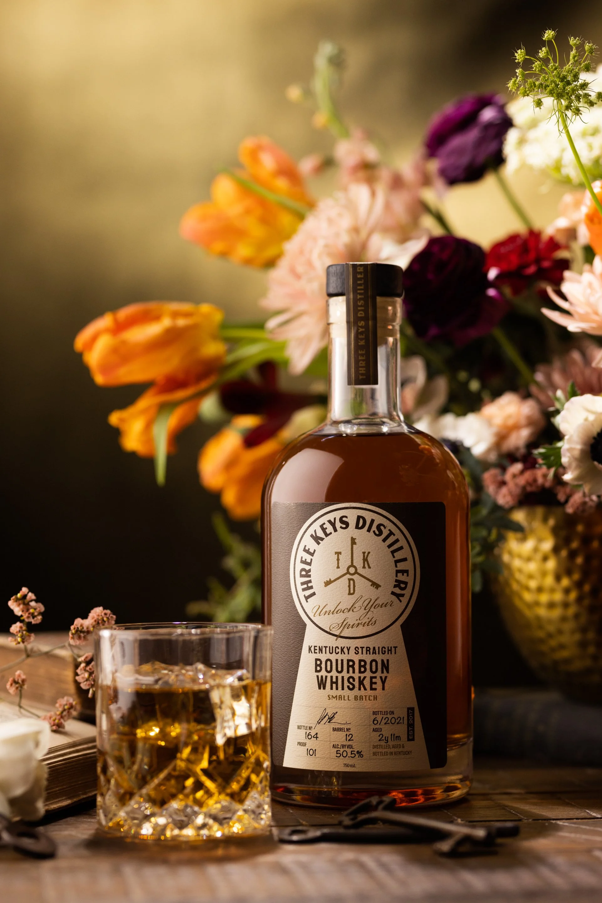 A bottle of Kentucky straight bourbon whiskey with a glass of whiskey on the rocks in front, placed on a wooden surface with a bouquet of colorful flowers in the background.