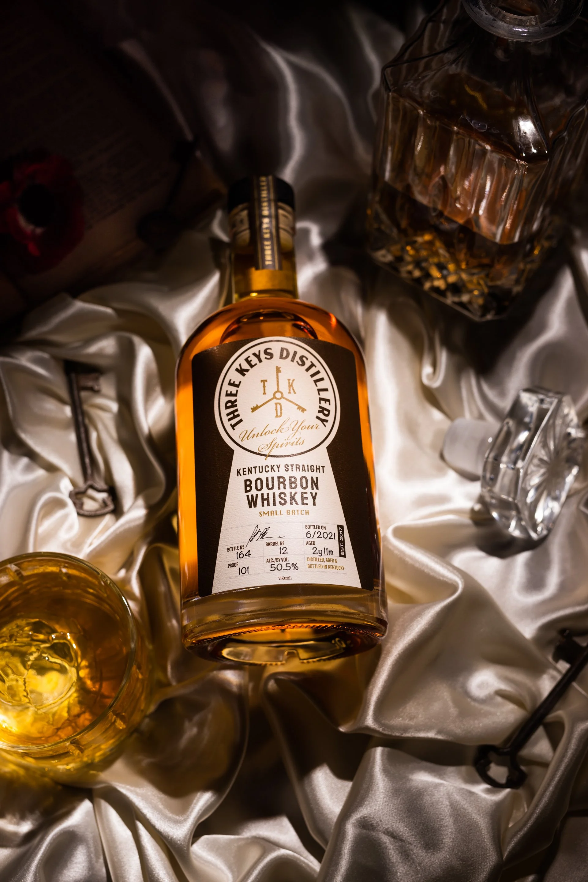 A bottle of Three Keys Distillery Kentucky Straight Bourbon Whiskey resting on light-colored satin fabric, with a glass of whiskey and additional glasses nearby.