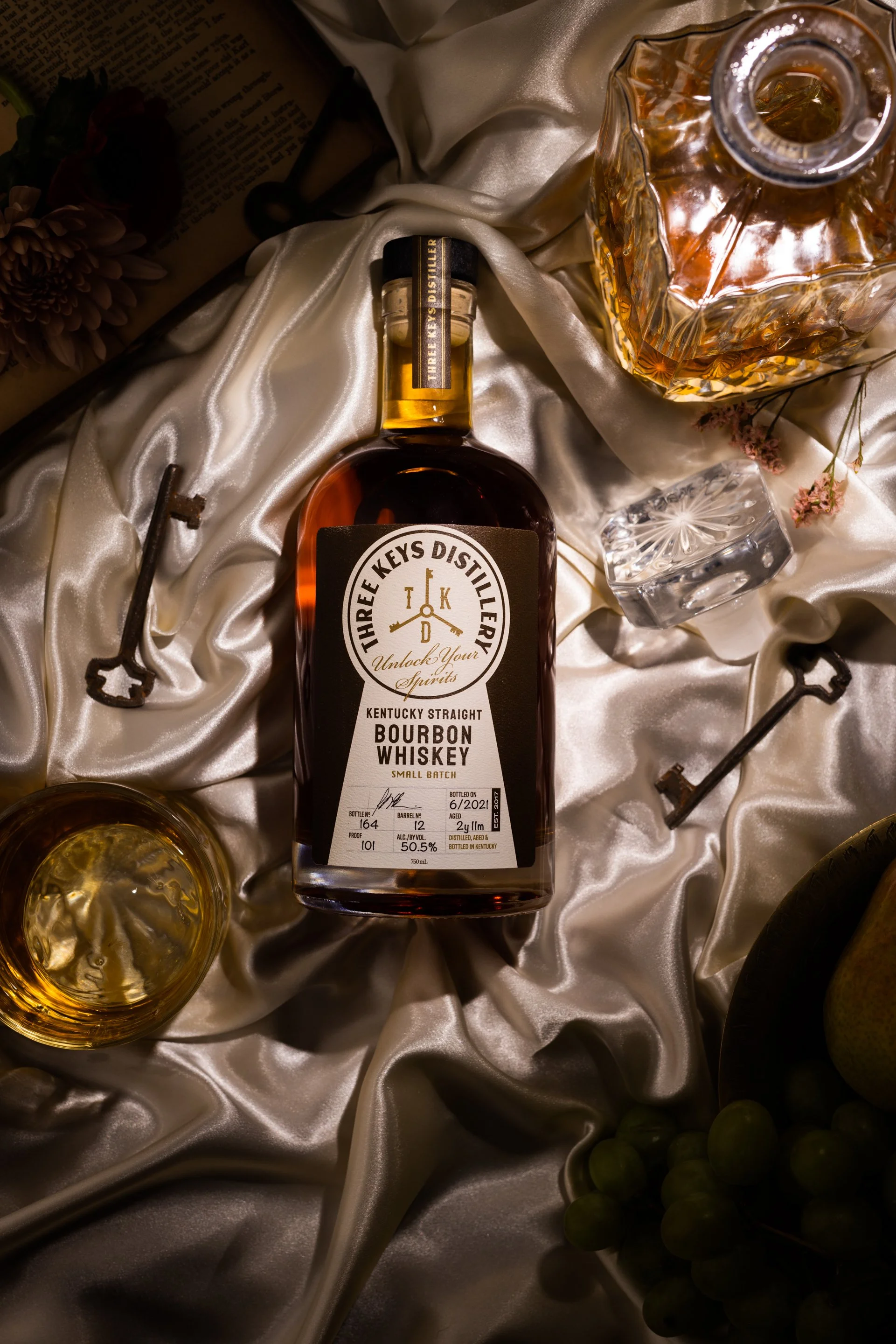 A bottle of Tharfe Keys distillery Kentucky straight bourbon whiskey surrounded by a glass of whiskey, a decanter, vintage keys, an ice cube, a bunch of grapes, and flowers on satin fabric.
