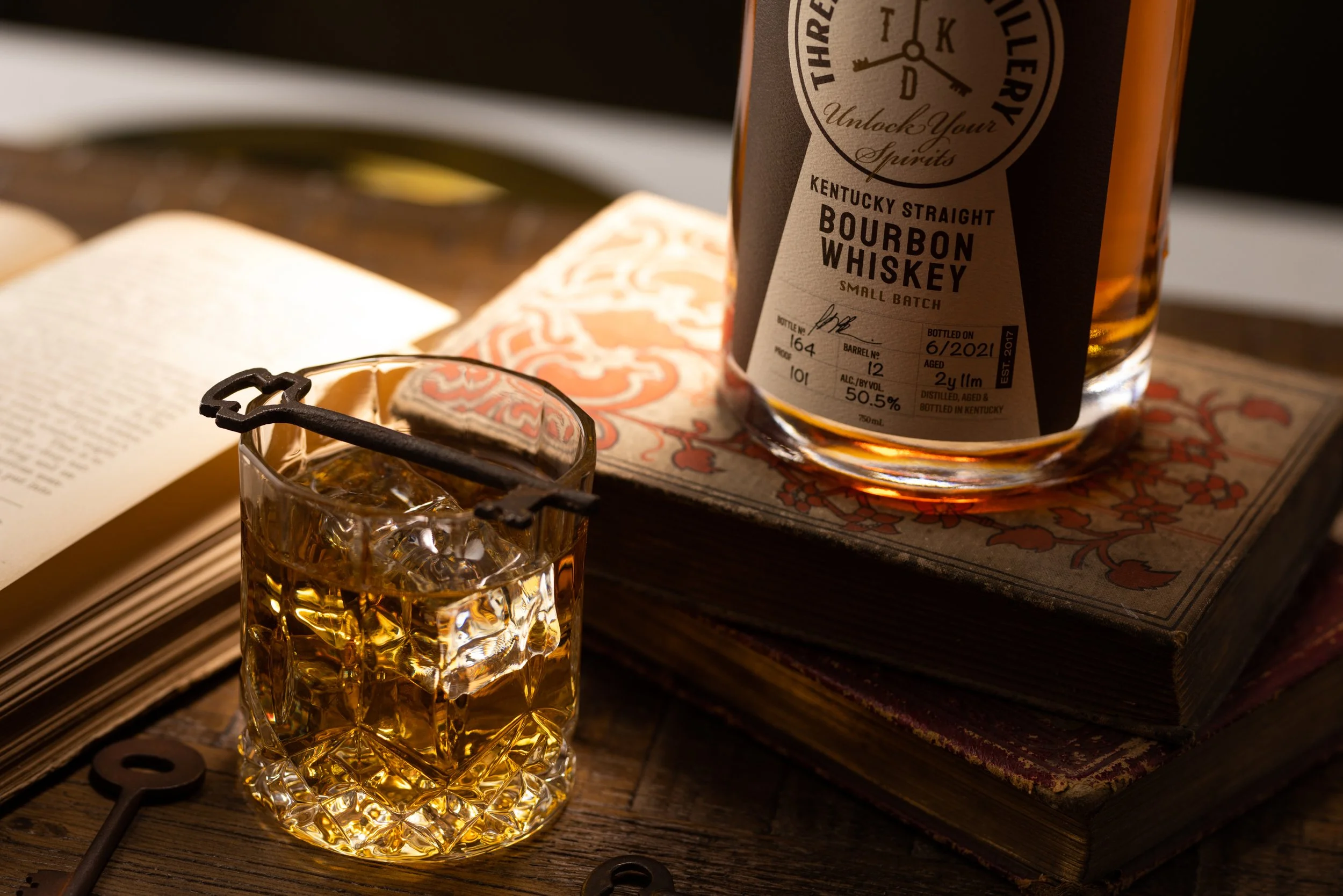 A glass of bourbon whiskey with ice cubes and a black key-shaped bottle opener on top, placed on a wooden surface next to an open book. A large bottle of Kentucky straight bourbon whiskey is partially visible, resting on a decorative book with vintag