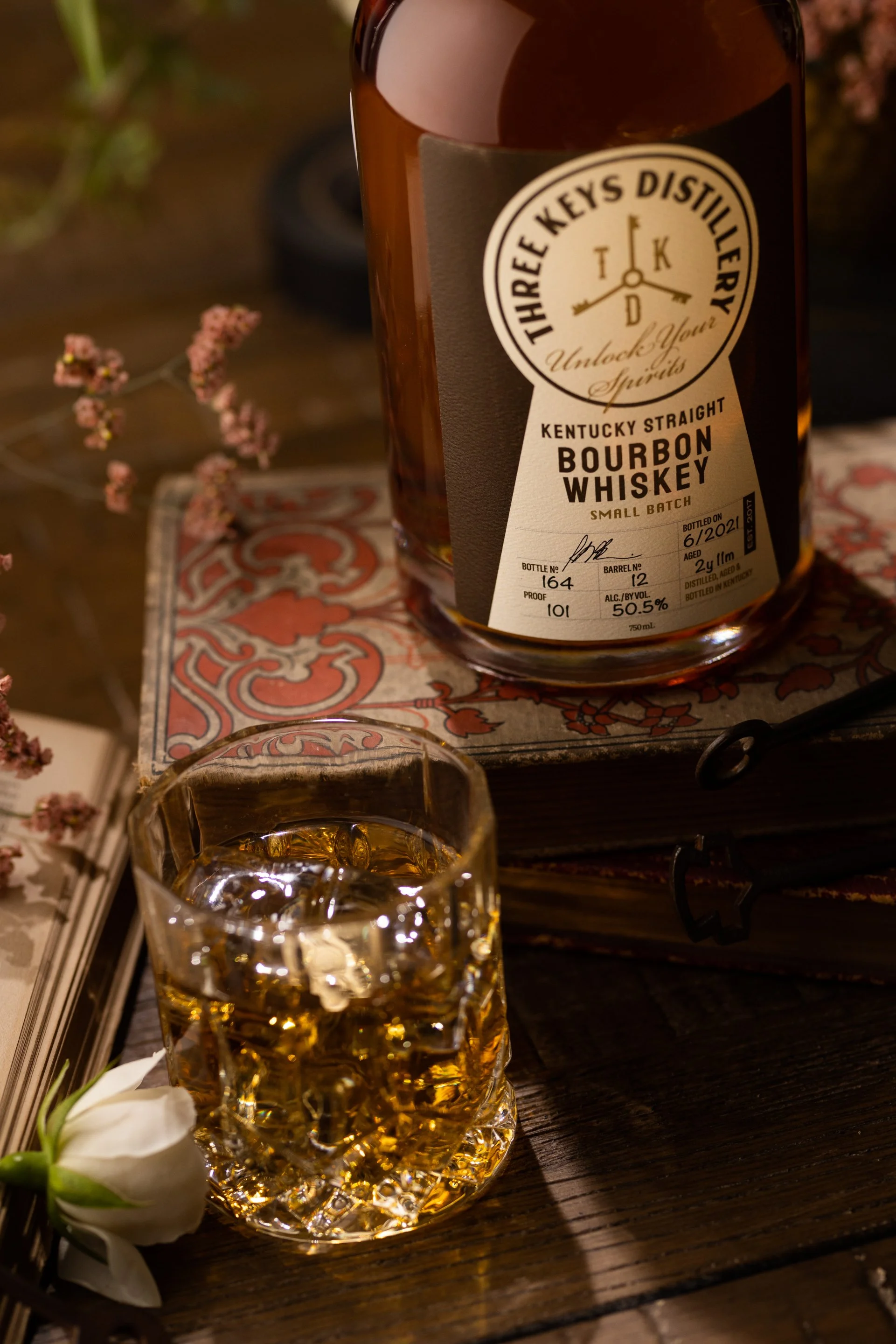 A bottle of Three Keys Distillery Kentucky Straight Bourbon Whiskey and a glass filled with whiskey and ice on a wooden surface, surrounded by flowers and decorative objects.