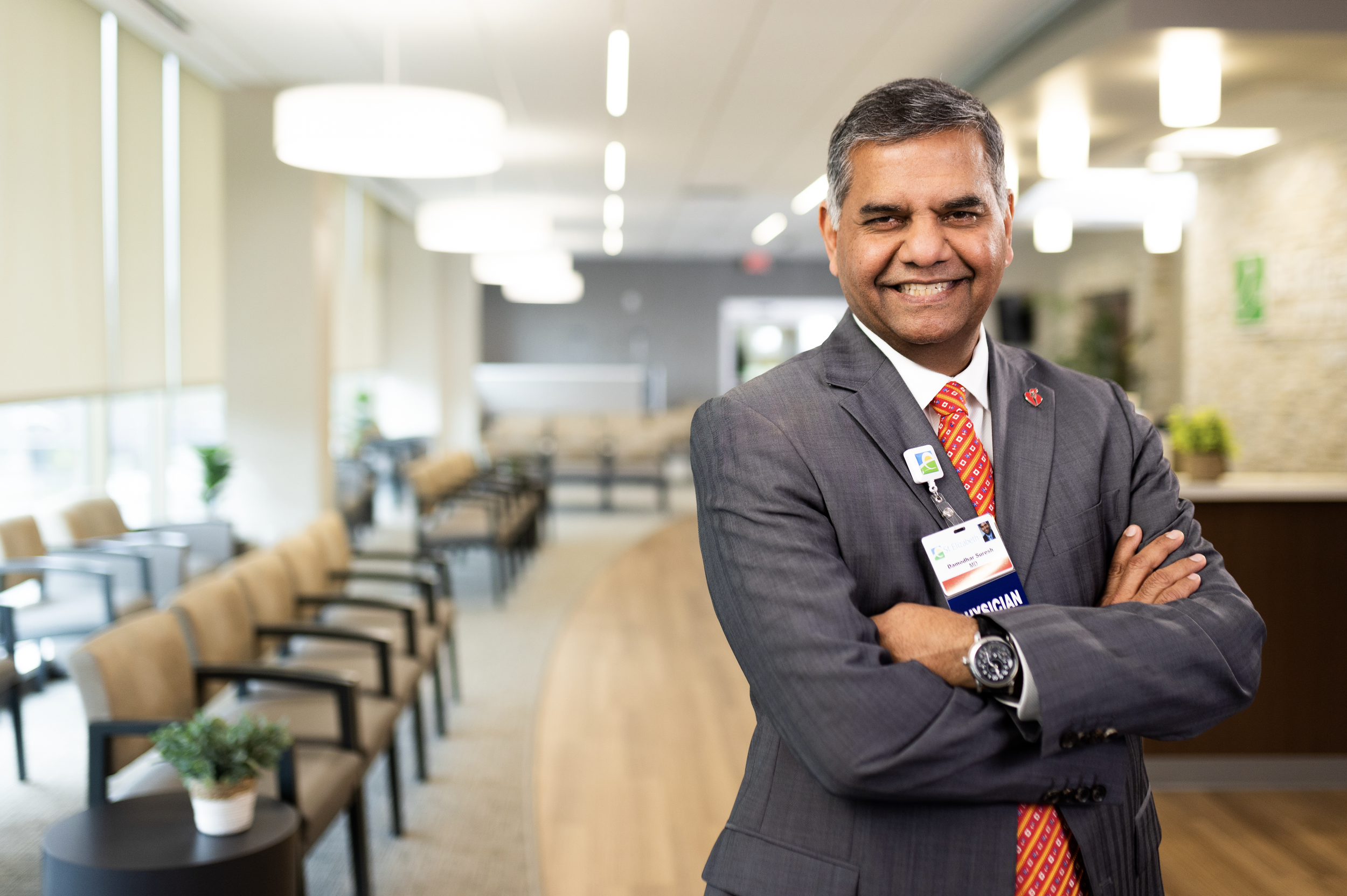 Smiling man in a business suit with a scrubs badge and a digital watch standing with arms crossed in a modern waiting area or office lobby.