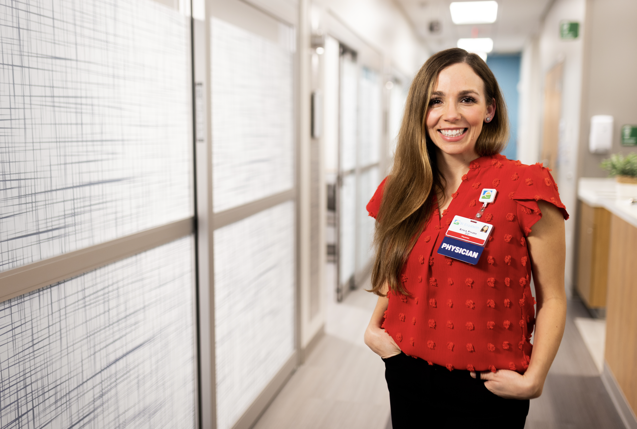 A smiling woman with long brown hair wearing a red top with ruffle details, a name badge, and a physician badge, standing in a hallway with white walls and lockers.