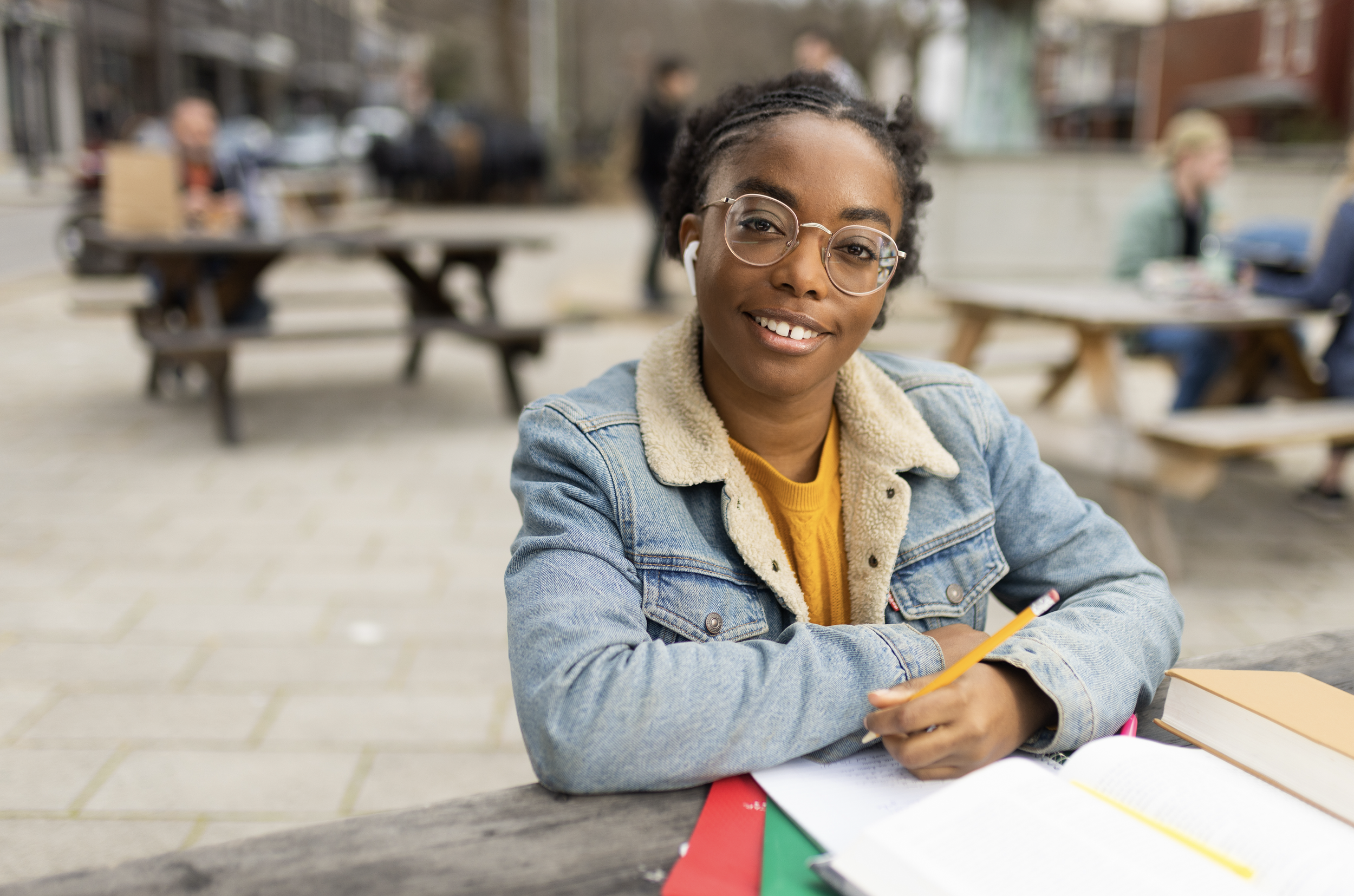 A young woman with glasses and a denim jacket, smiling at the camera, sitting at an outdoor table with books and a pencil, with people in the background.