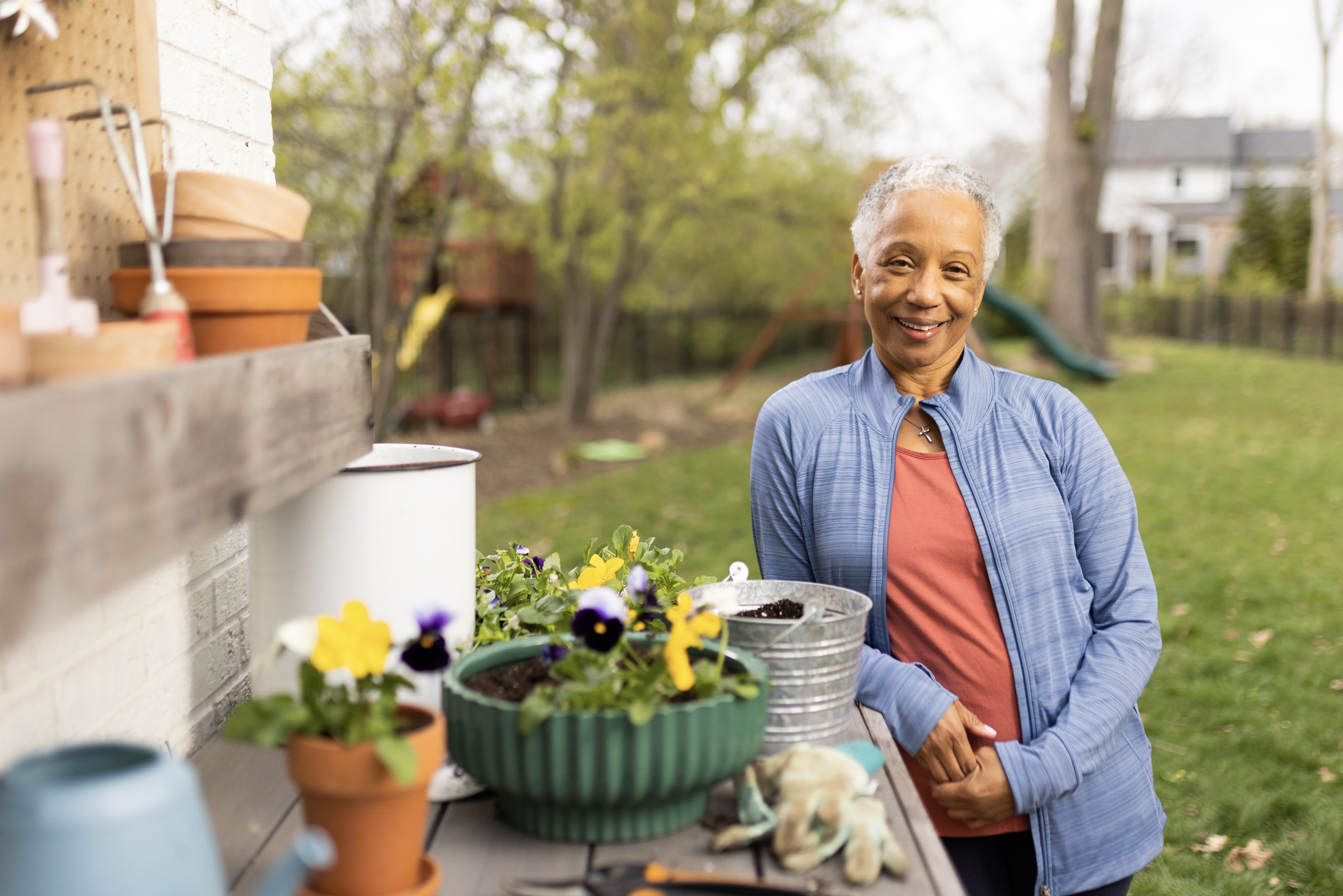 A smiling elderly woman in a blue jacket and orange shirt standing in a backyard garden, surrounded by potted flowers and gardening tools.